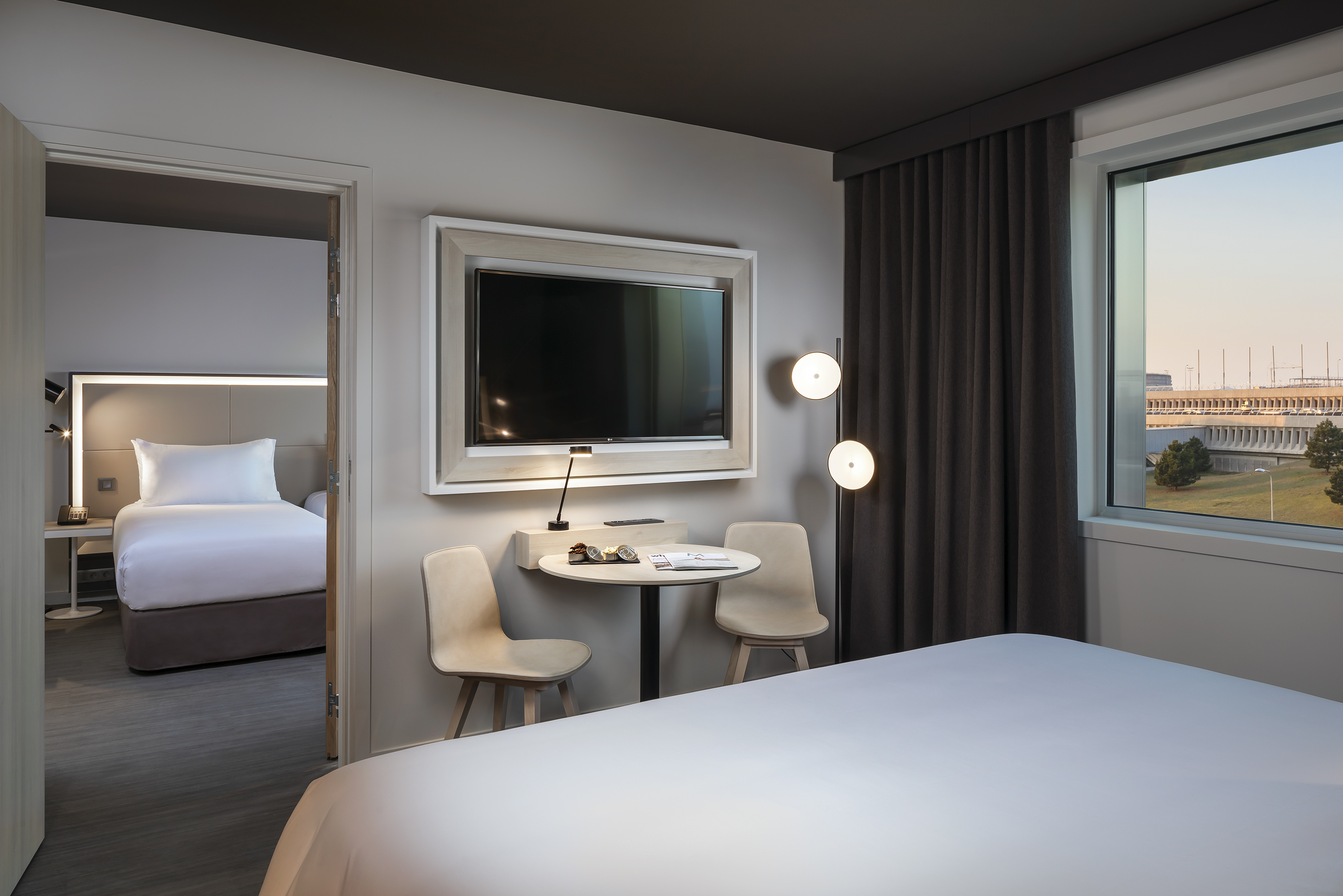 Modern hotel room with two beds, TV, table, and airport window view.