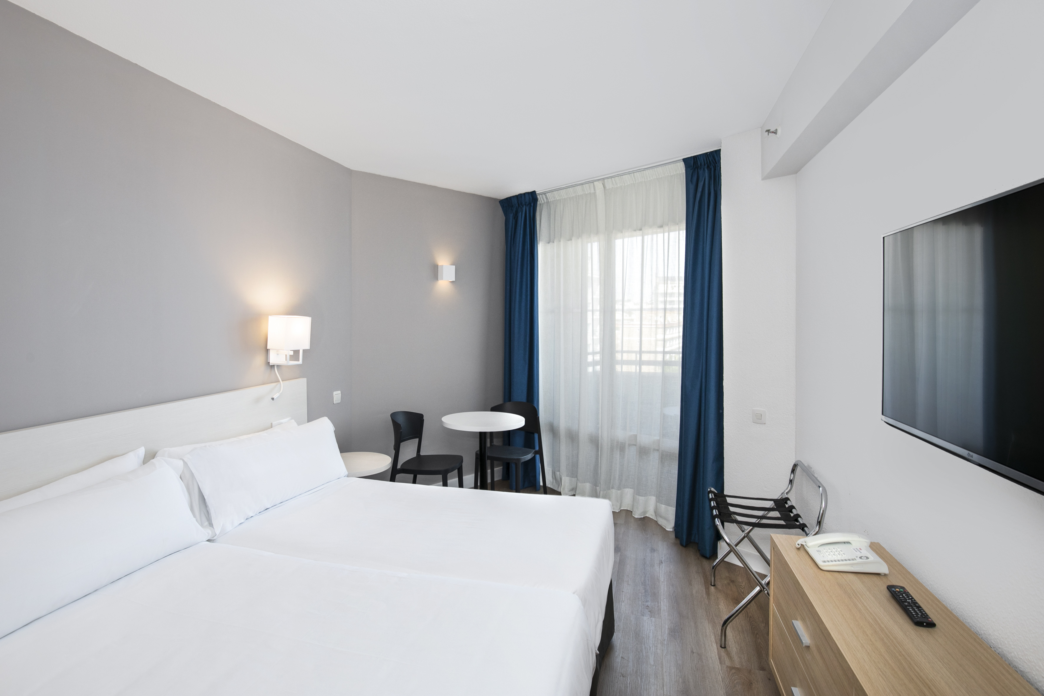 Modern hotel room with twin beds, blue curtains, TV, and table.
