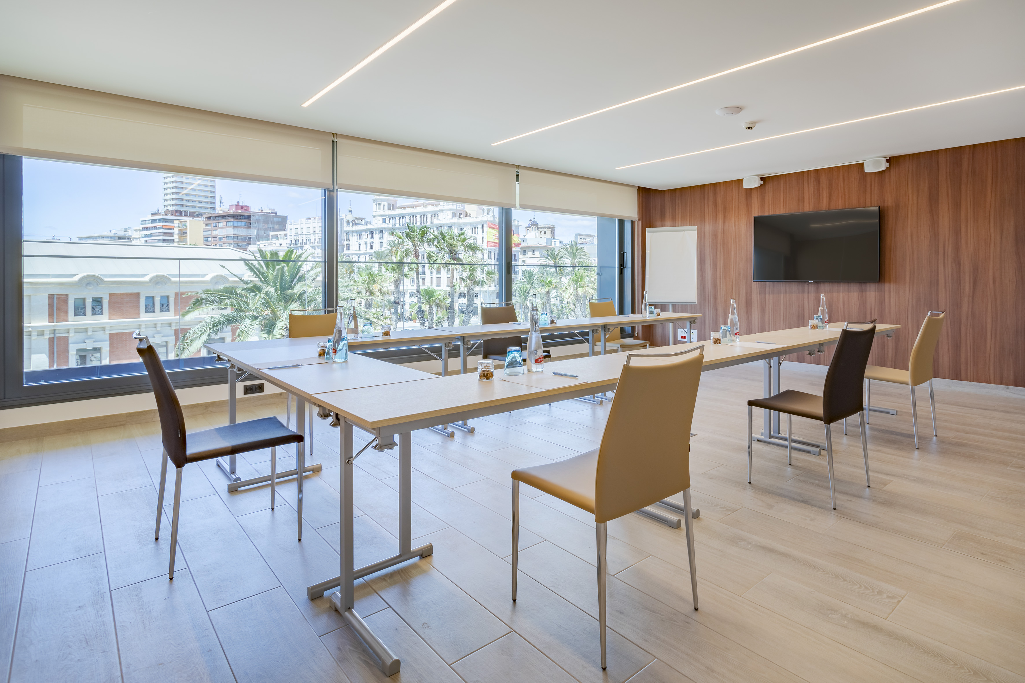 Modern meeting room with city view, U-shaped tables, large TV, and whiteboard.