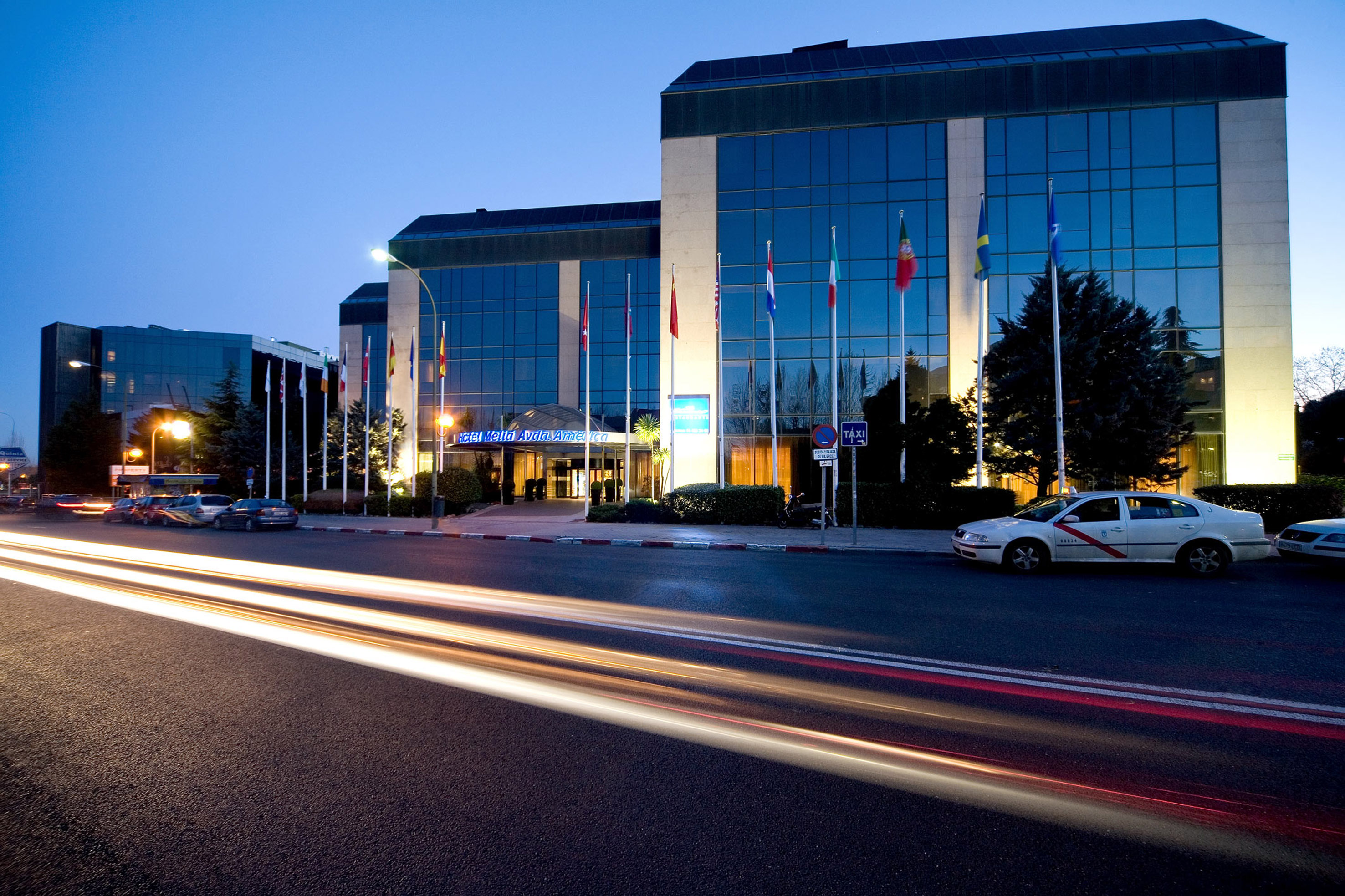 Hotel Meliá Avda América at dusk, modern glass building, flags, and car light trails.