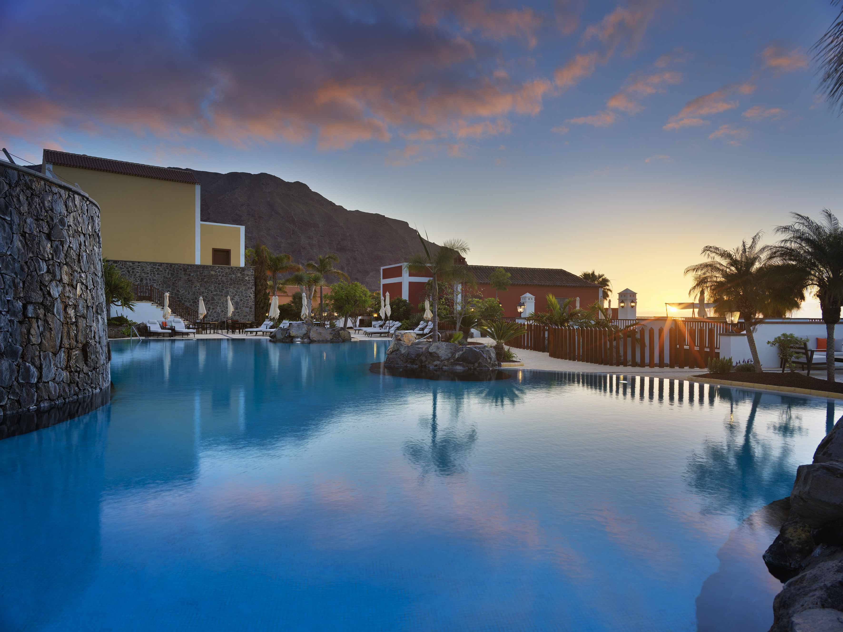 Tropical resort pool reflecting vibrant sunset sky, palm trees, and mountains.