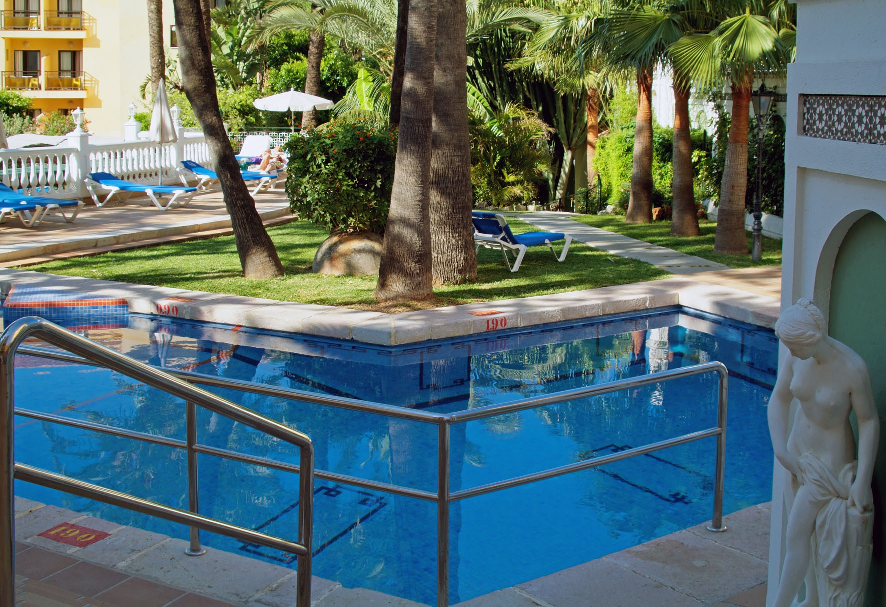 Resort swimming pool with palm trees, blue sun loungers, and a statue.