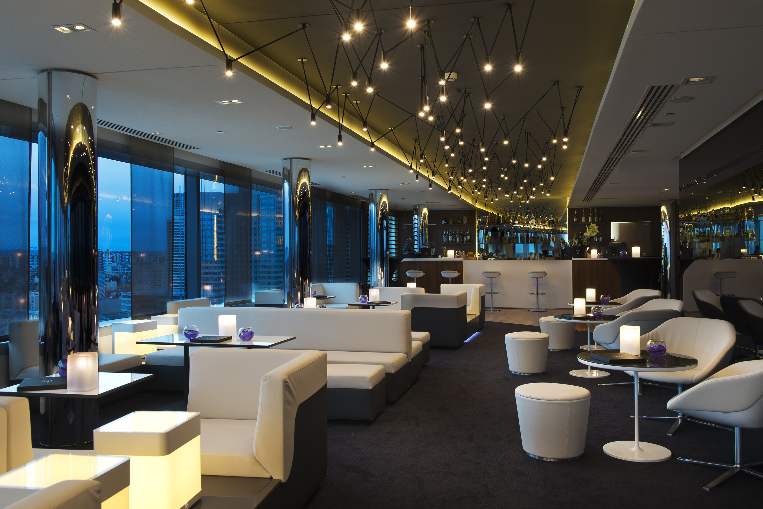 Modern bar lounge with city skyline views, white seating, and decorative lighting.