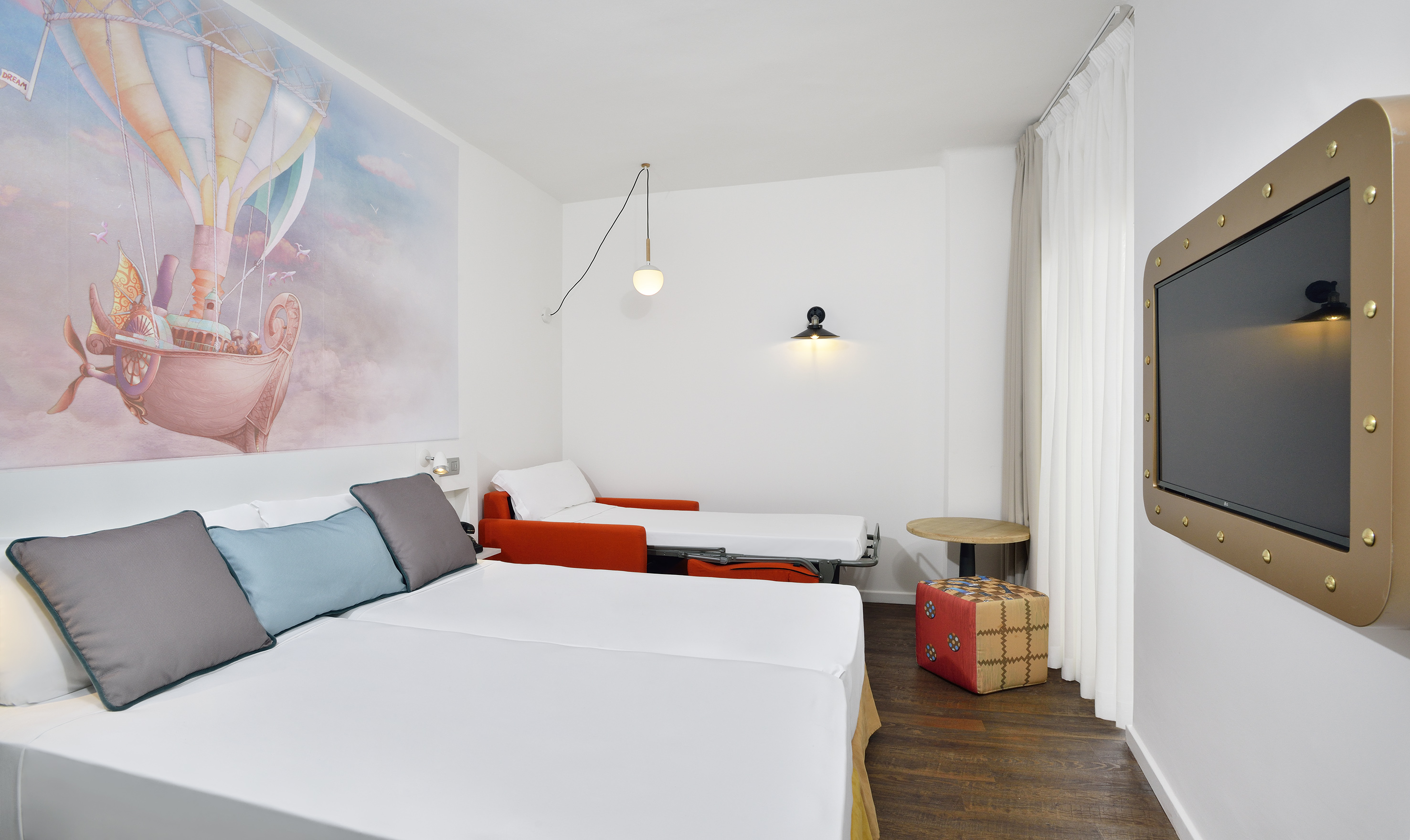 Hotel room with two beds, hot air balloon mural, sofa bed, and gold-framed TV.