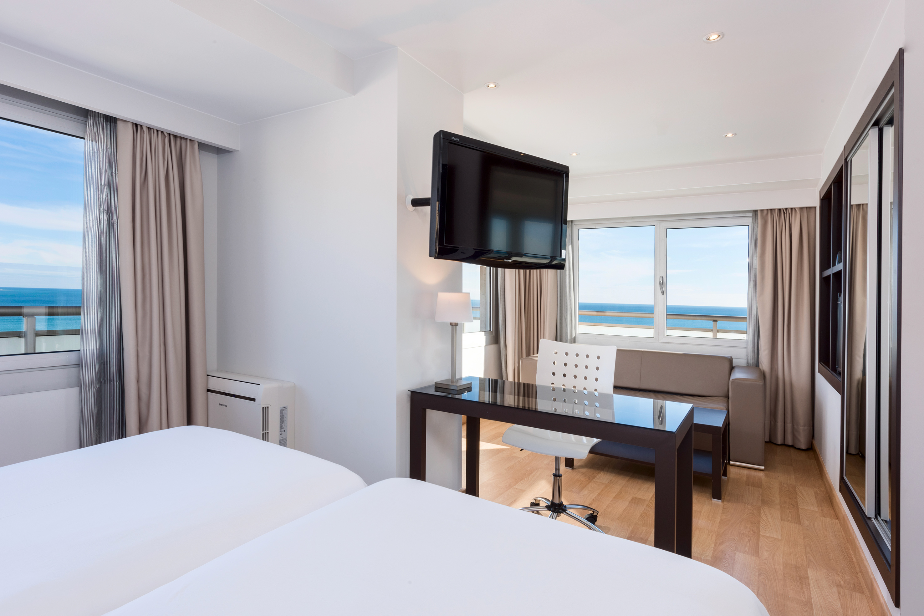 Modern hotel room with ocean view, twin beds, desk, and wall TV.