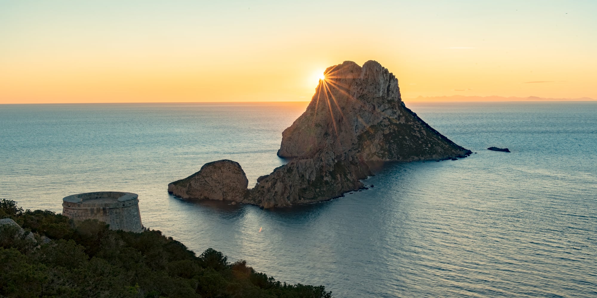 Atardecer sobre Es Vedrà en Ibiza con rayos de sol, mar y torre antigua.
