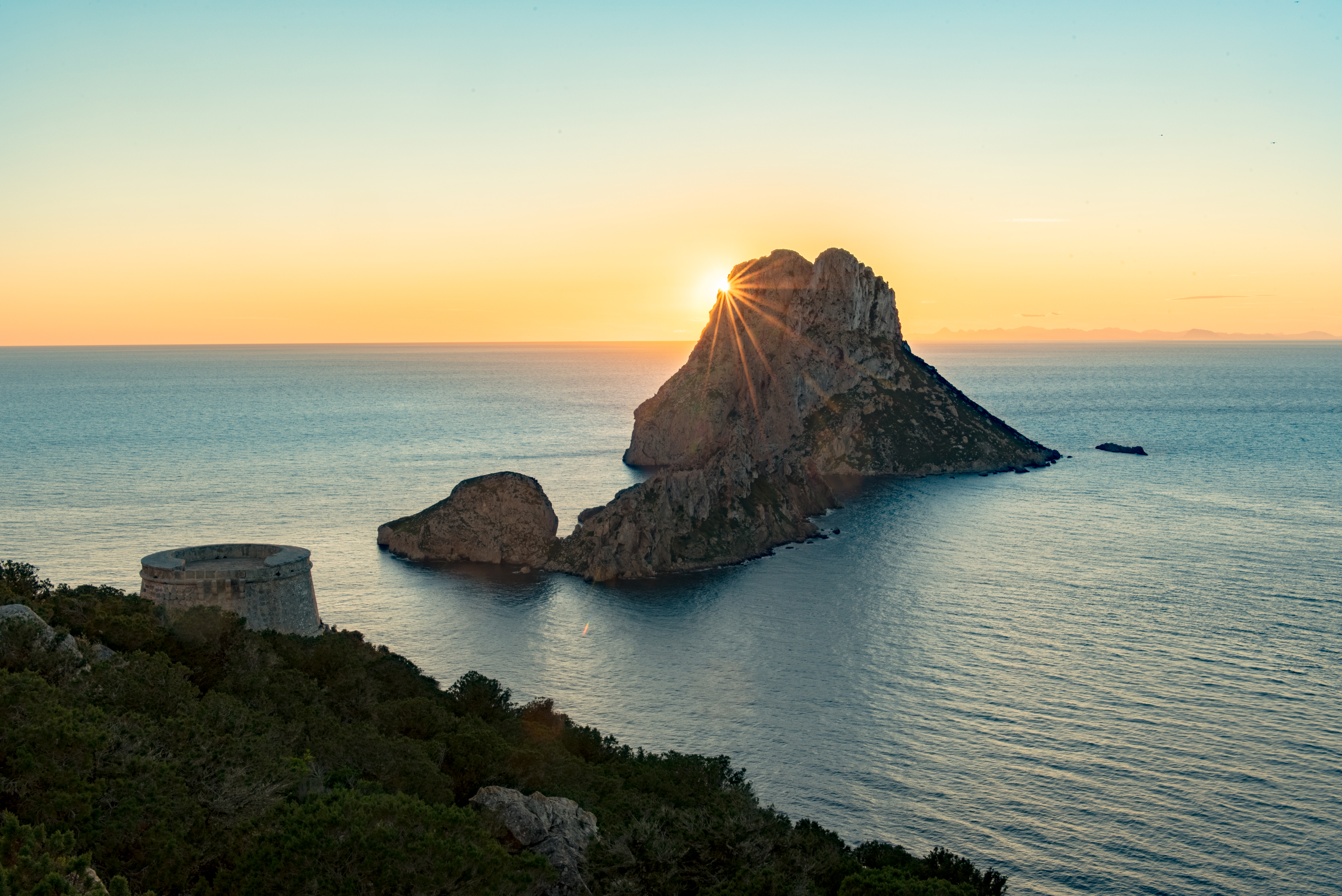 Atardecer sobre Es Vedrà en Ibiza con rayos de sol, mar y torre antigua.