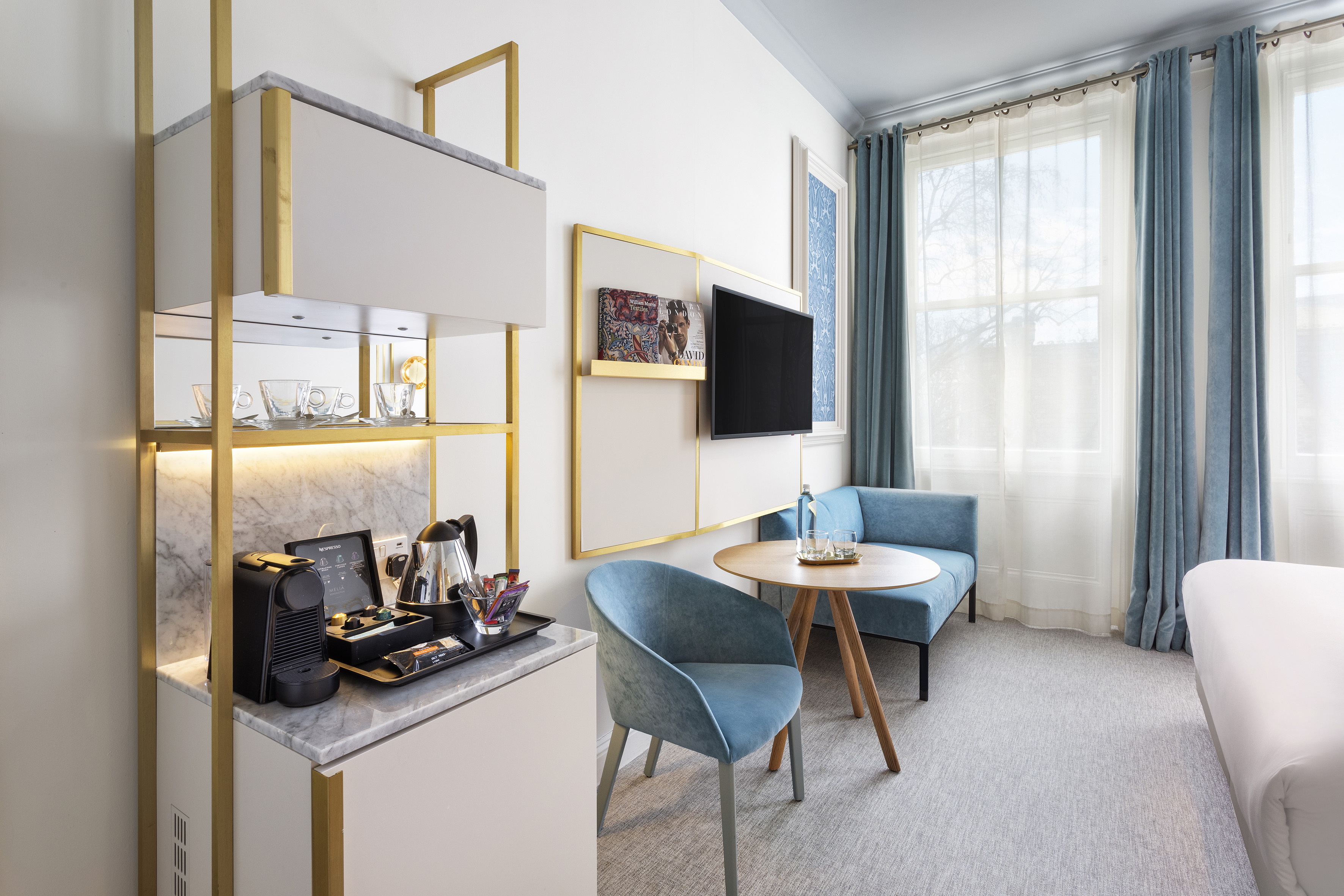 Elegant hotel room: Nespresso coffee bar, blue seating, TV, and bright windows.