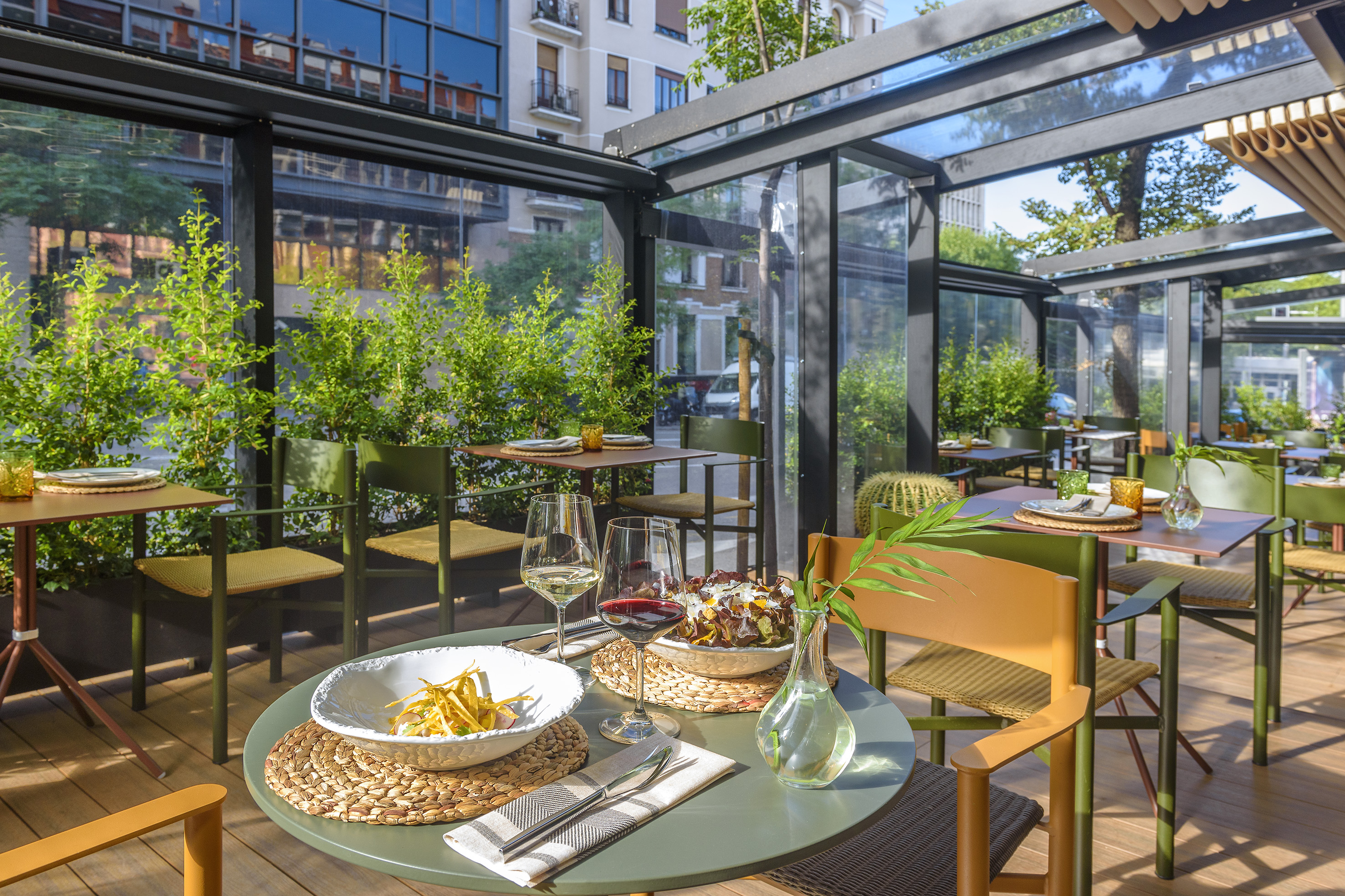 Sunny outdoor restaurant terrace with glass roof, set tables, wine, and lush greenery.