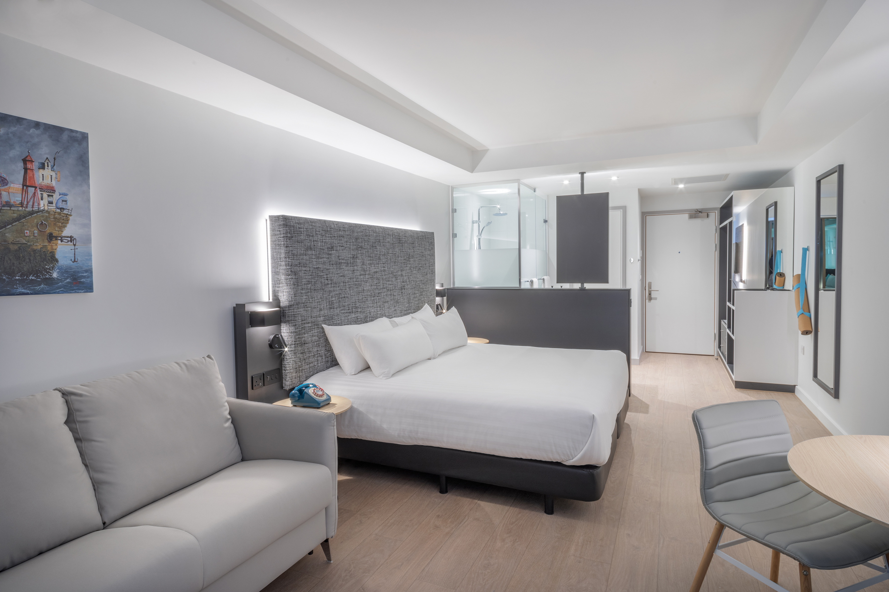 Modern hotel room with king bed, gray sofa, wood floor, and glass shower.