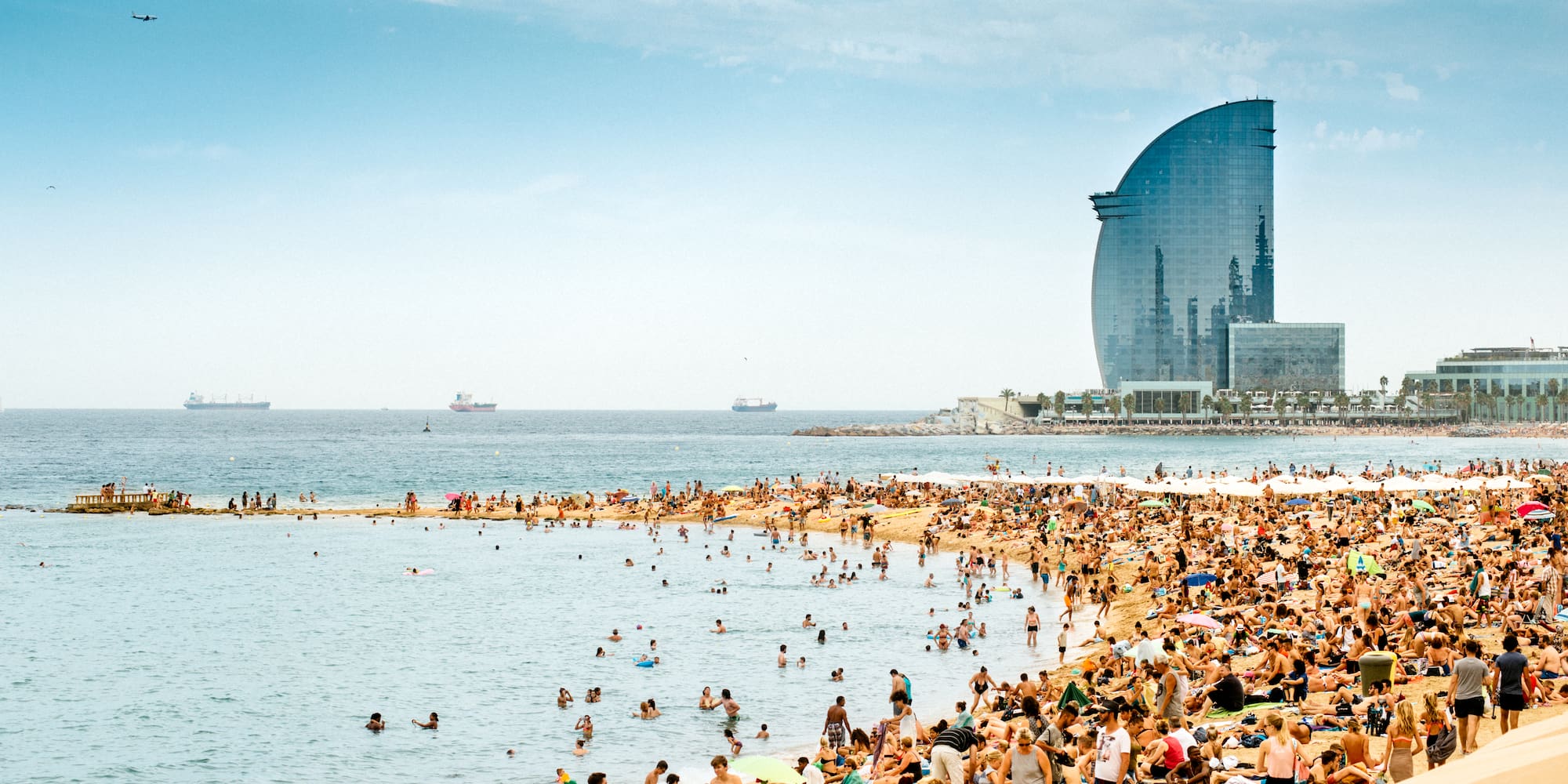 Playa Barceloneta concurrida, gente nadando, Hotel W Barcelona al fondo.