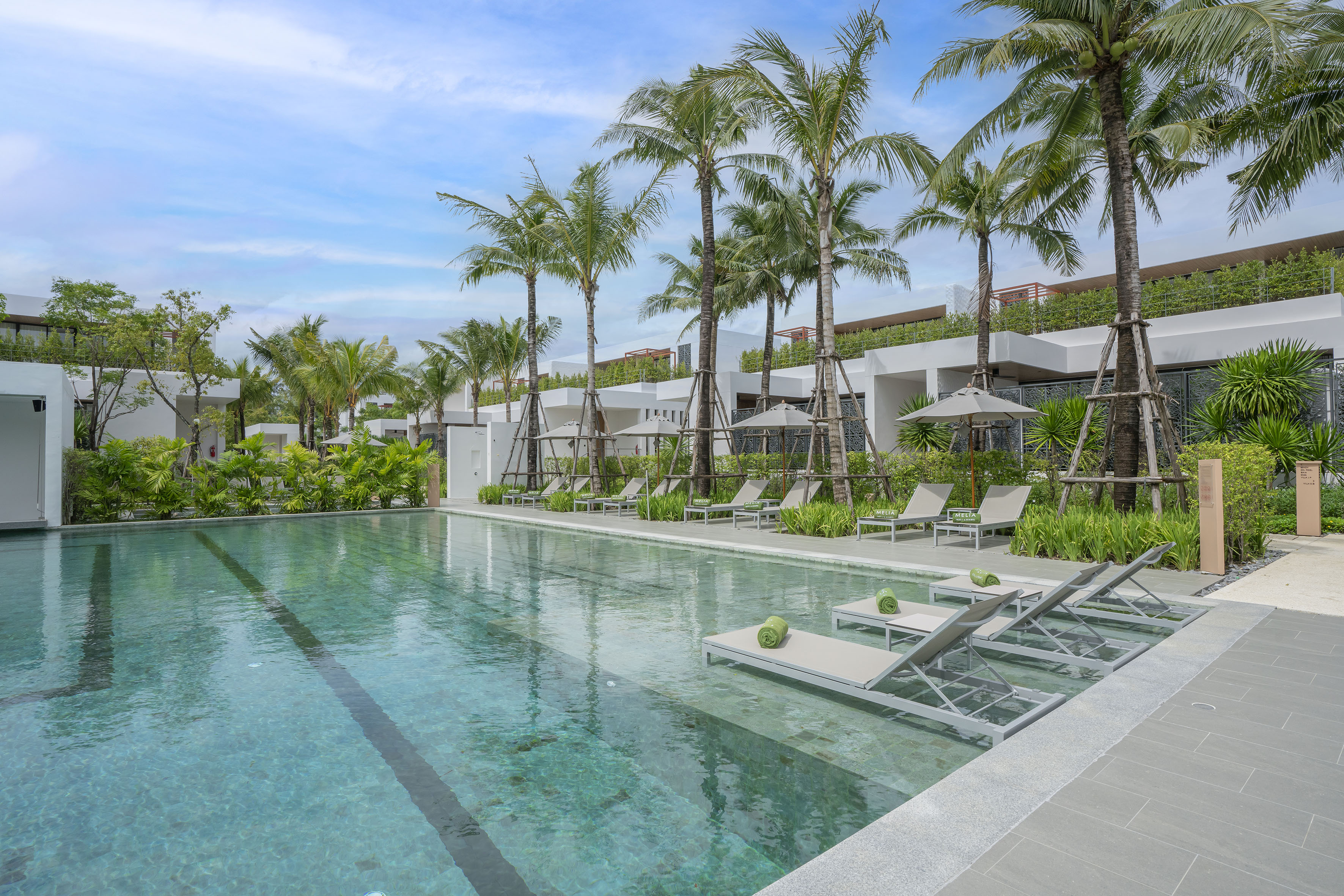 Resort swimming pool lined with palm trees, sun loungers, and modern buildings.