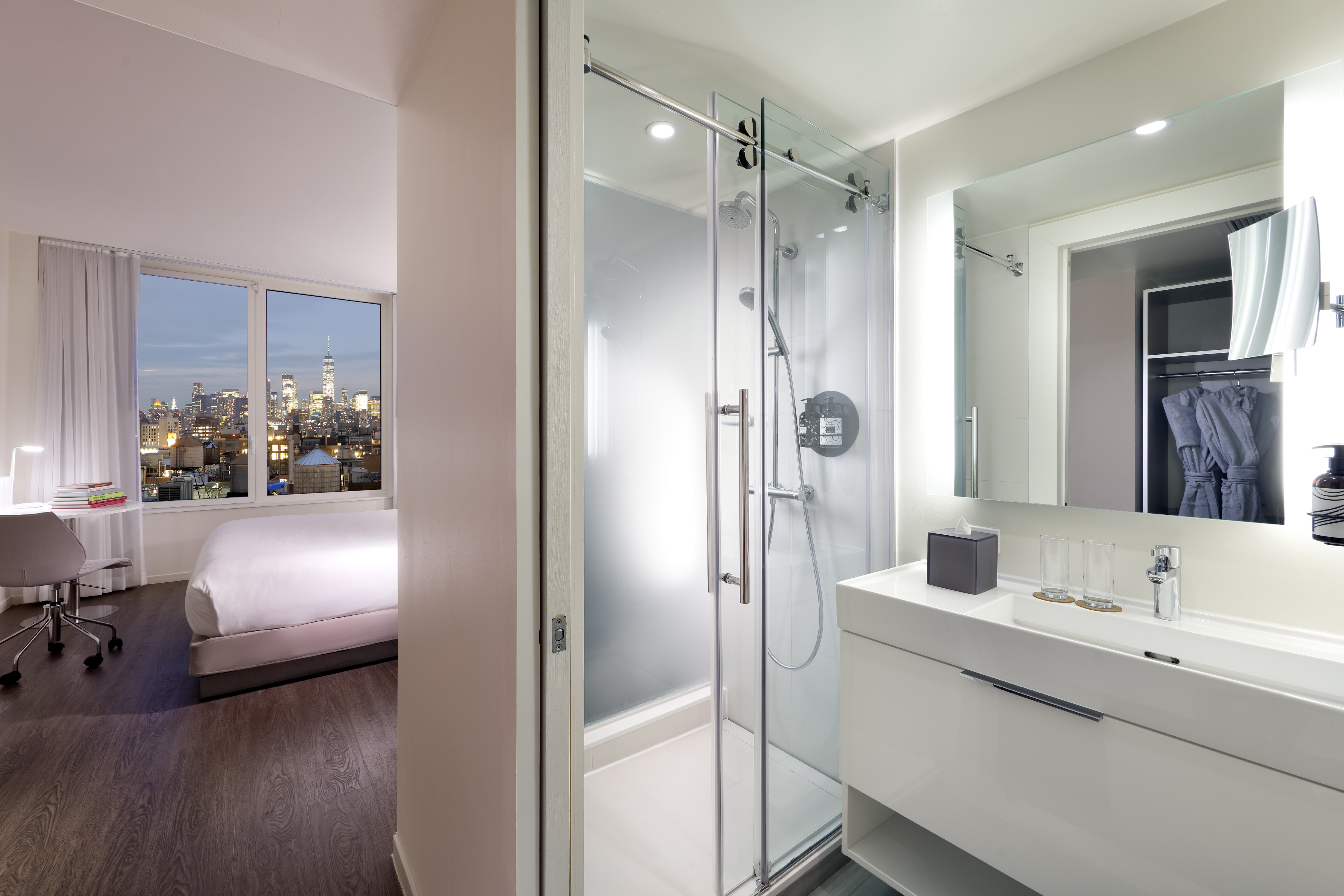 Modern hotel room with city skyline view, open-concept bathroom and glass shower.