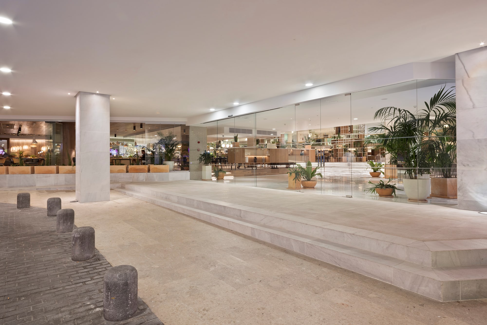 Modern hotel entrance with glass facade, bright lobby, stone steps and bollards.