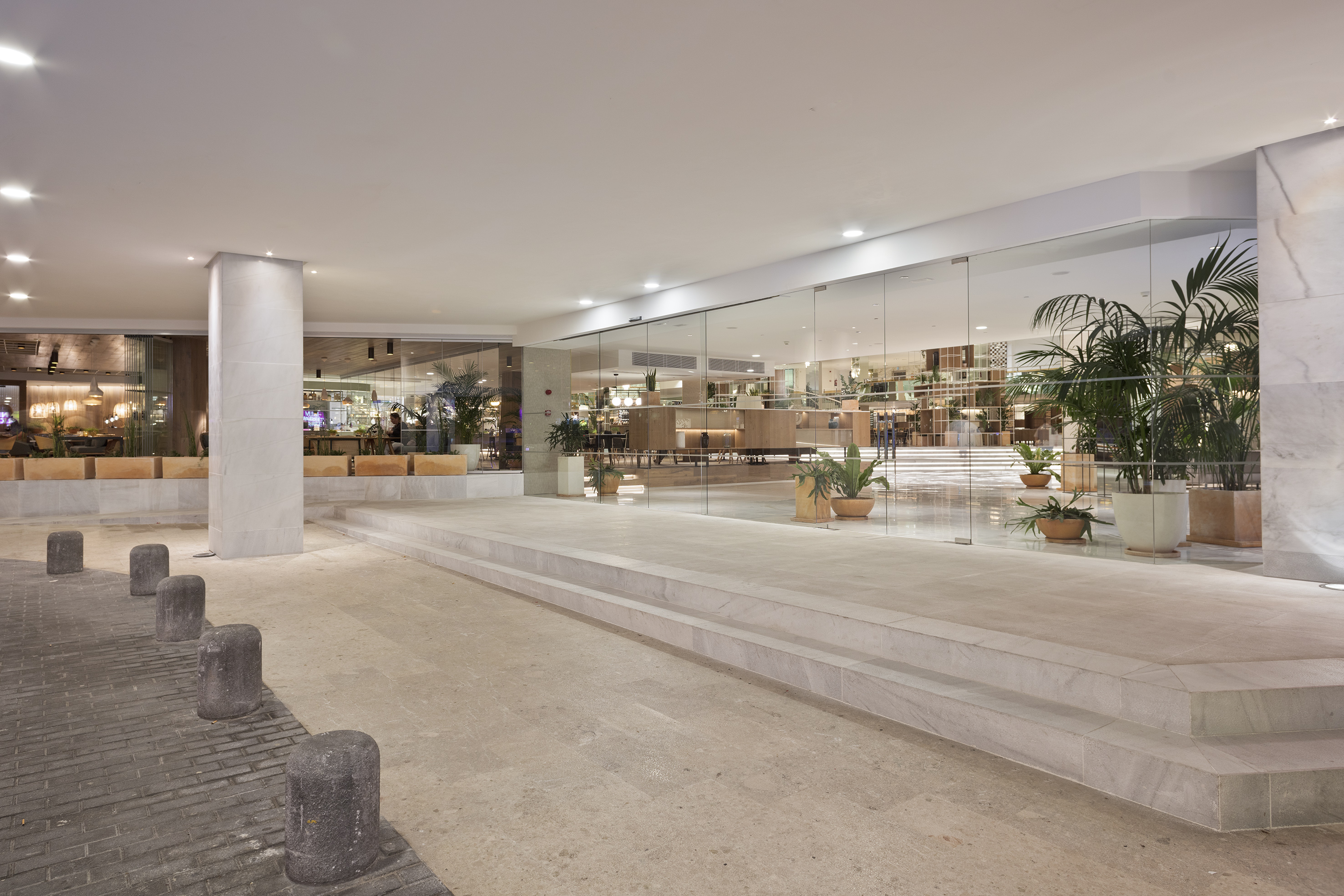 Modern hotel entrance with glass facade, bright lobby, stone steps and bollards.
