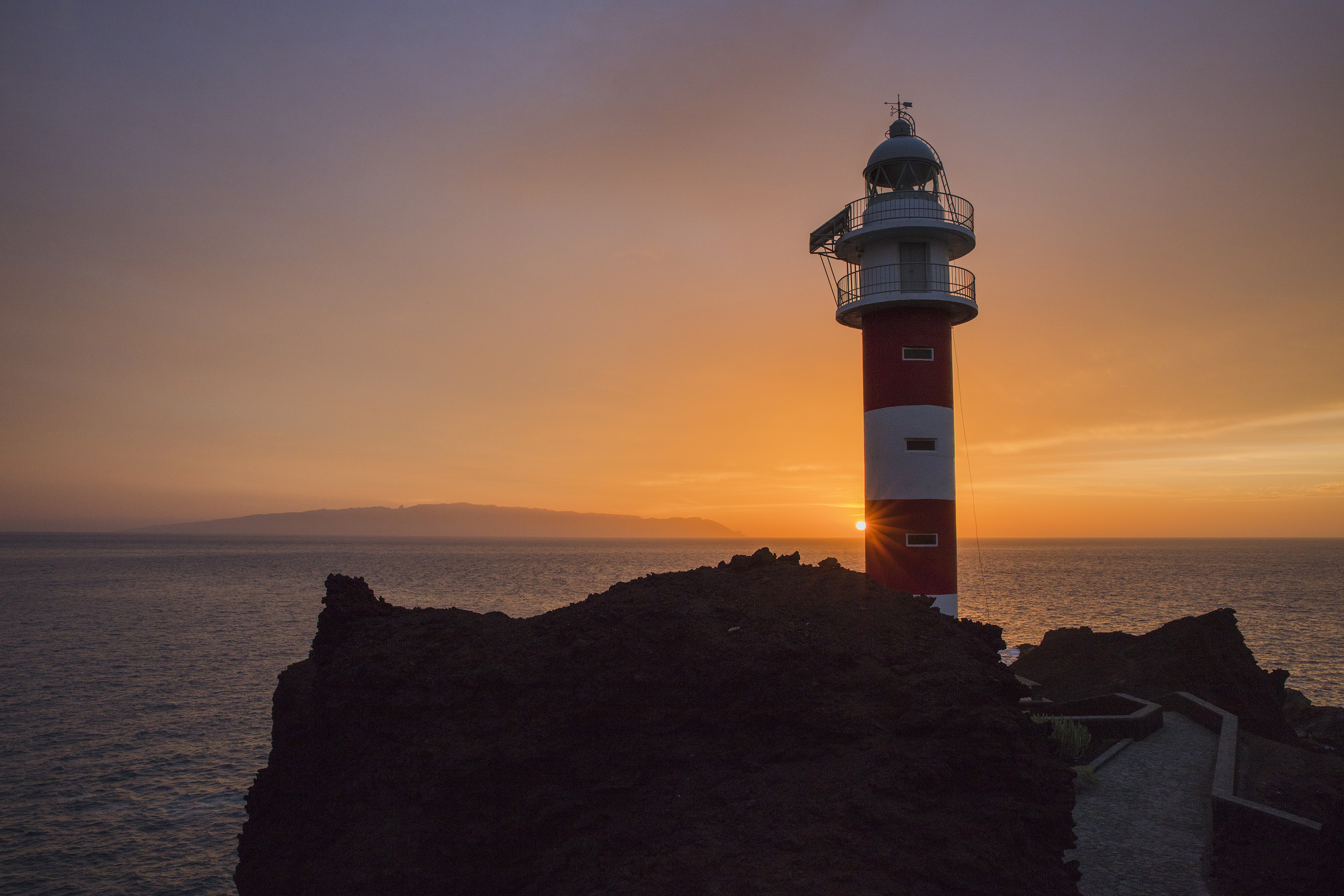 Red and white lighthouse on rocky coast with sun setting over the ocean.