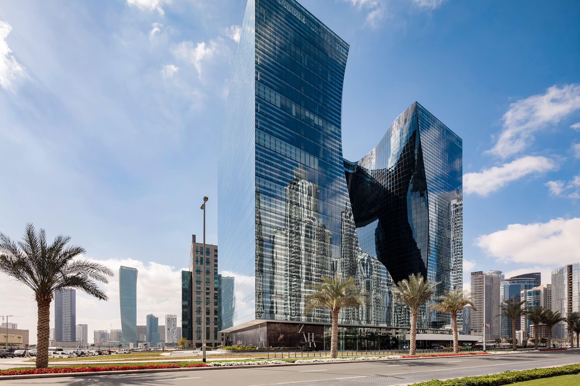 Modern glass skyscraper with curved design, city reflections, palm trees, blue sky.