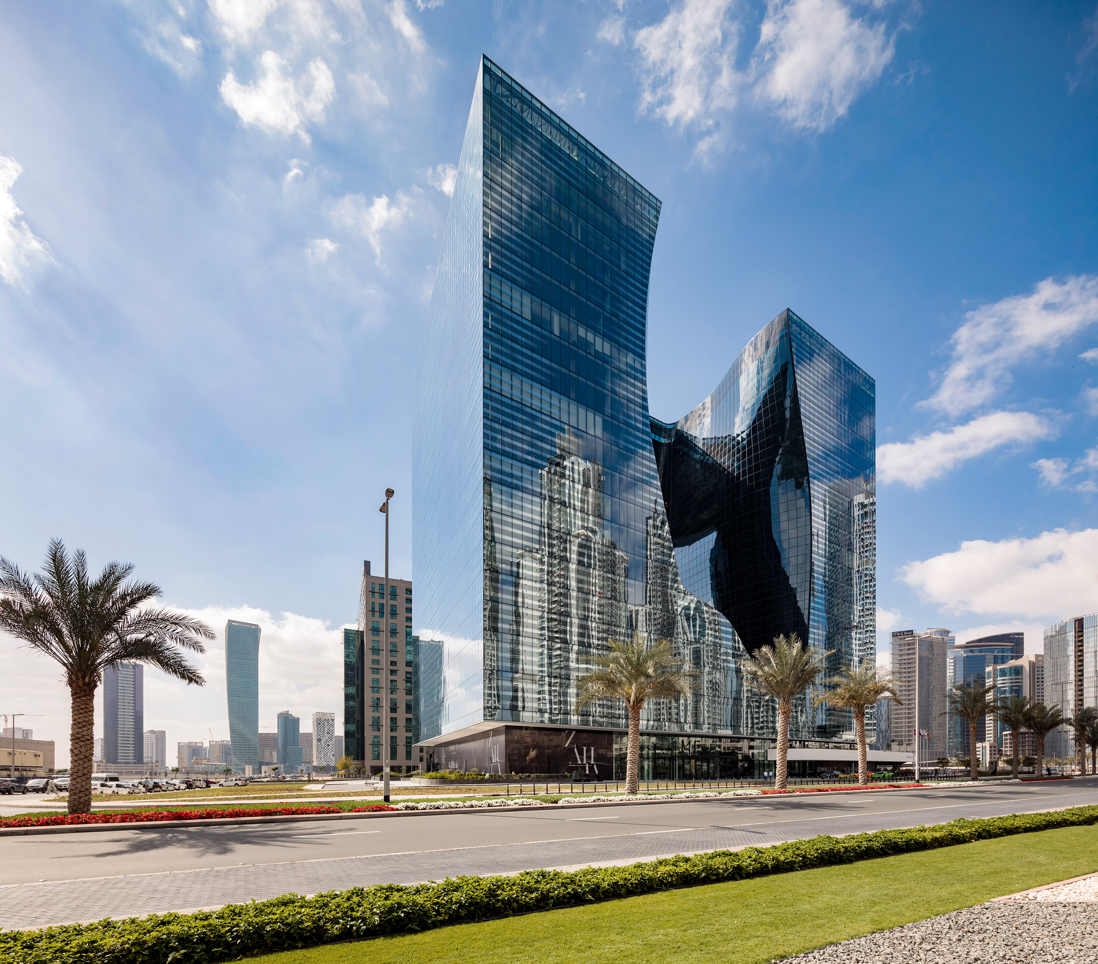 Modern glass skyscraper with curved design, city reflections, palm trees, blue sky.