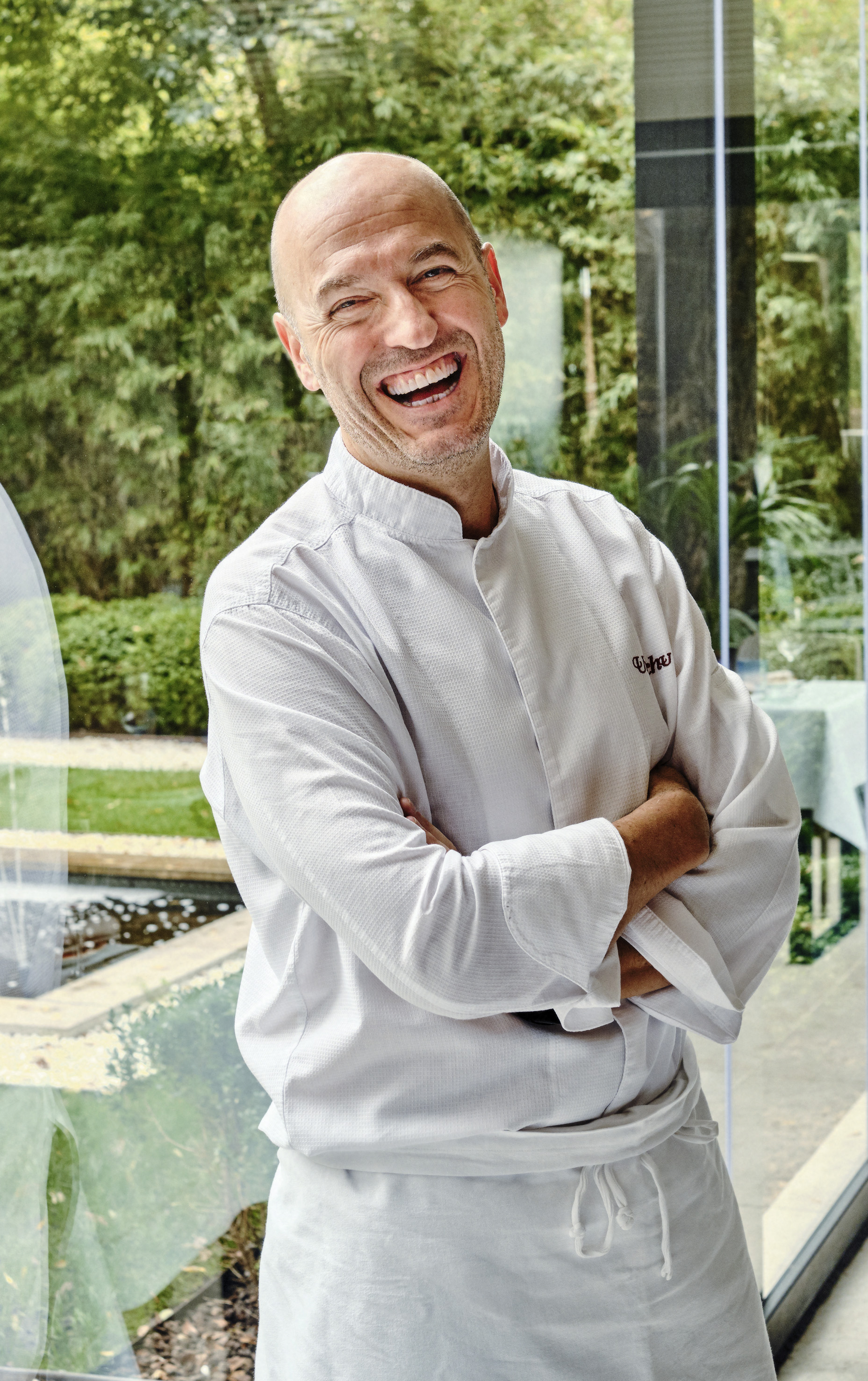 Bald chef in white uniform laughing, arms crossed, with green outdoor view.