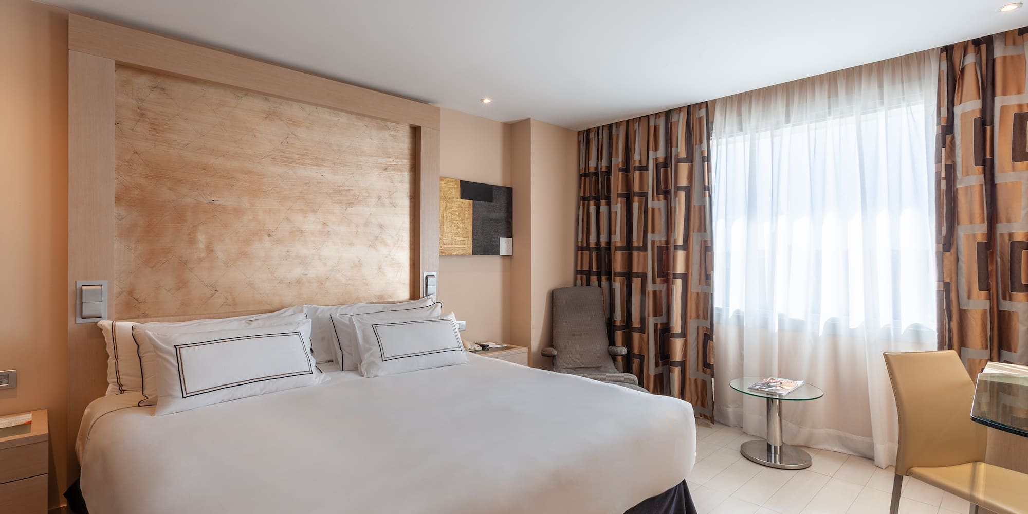 Modern hotel room with king bed, wood headboard, patterned curtains, and desk.