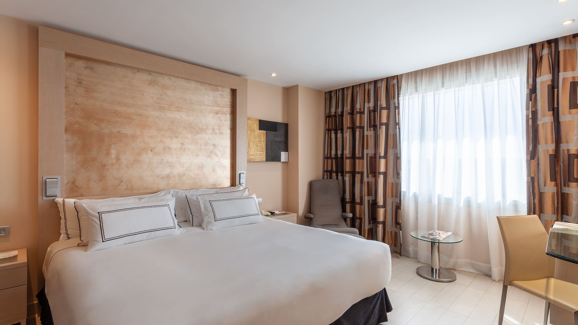 Modern hotel room with king bed, wood headboard, patterned curtains, and desk.