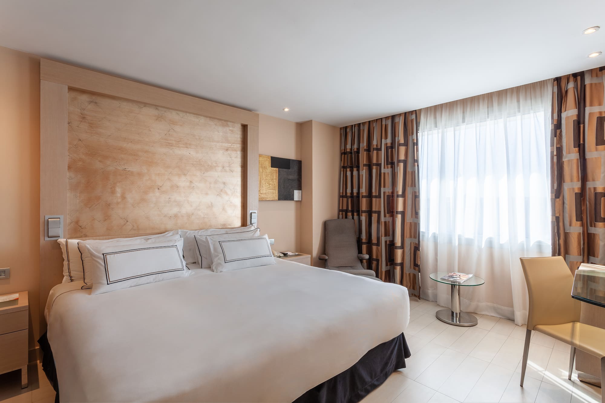 Modern hotel room with king bed, wood headboard, patterned curtains, and desk.