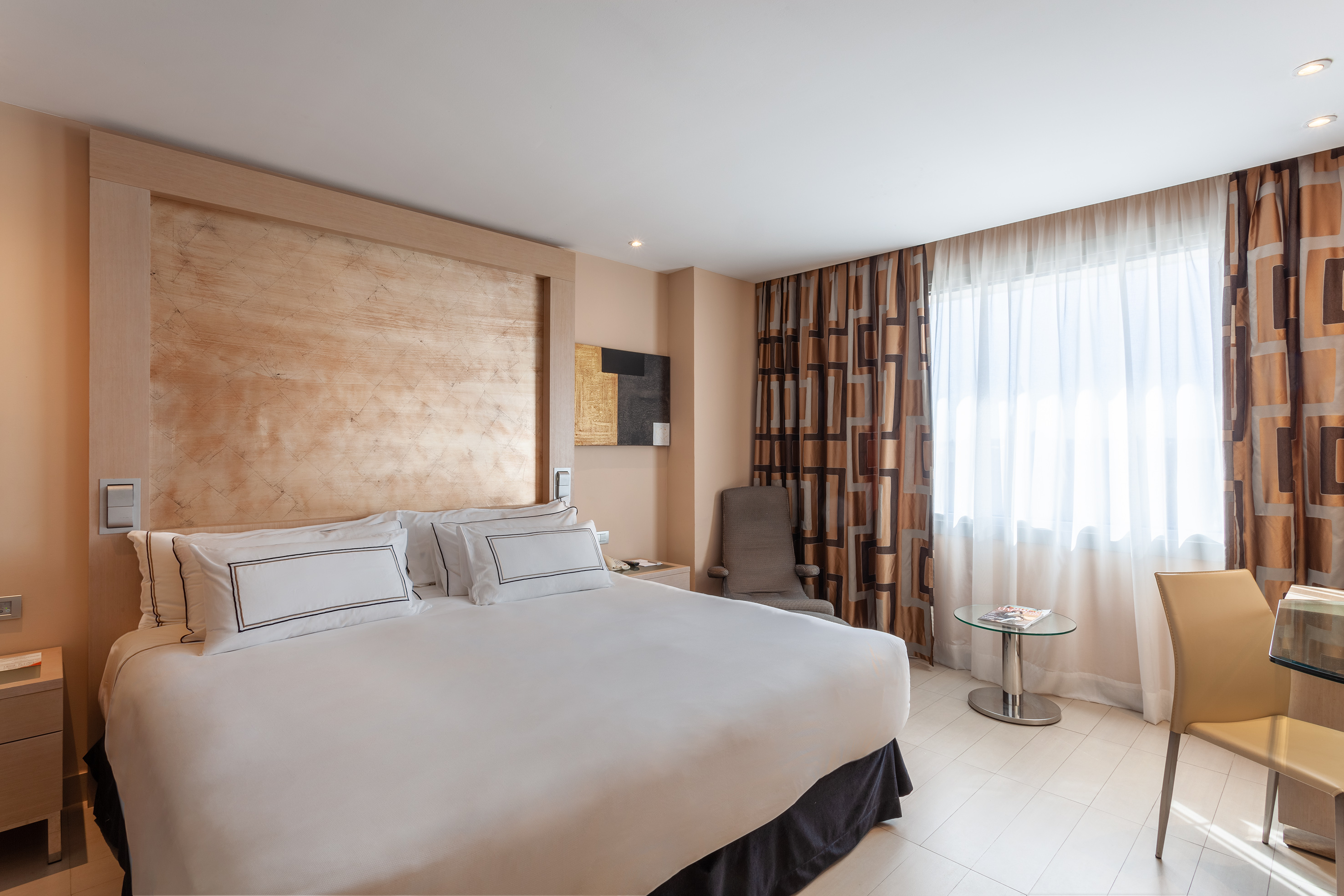 Modern hotel room with king bed, wood headboard, patterned curtains, and desk.