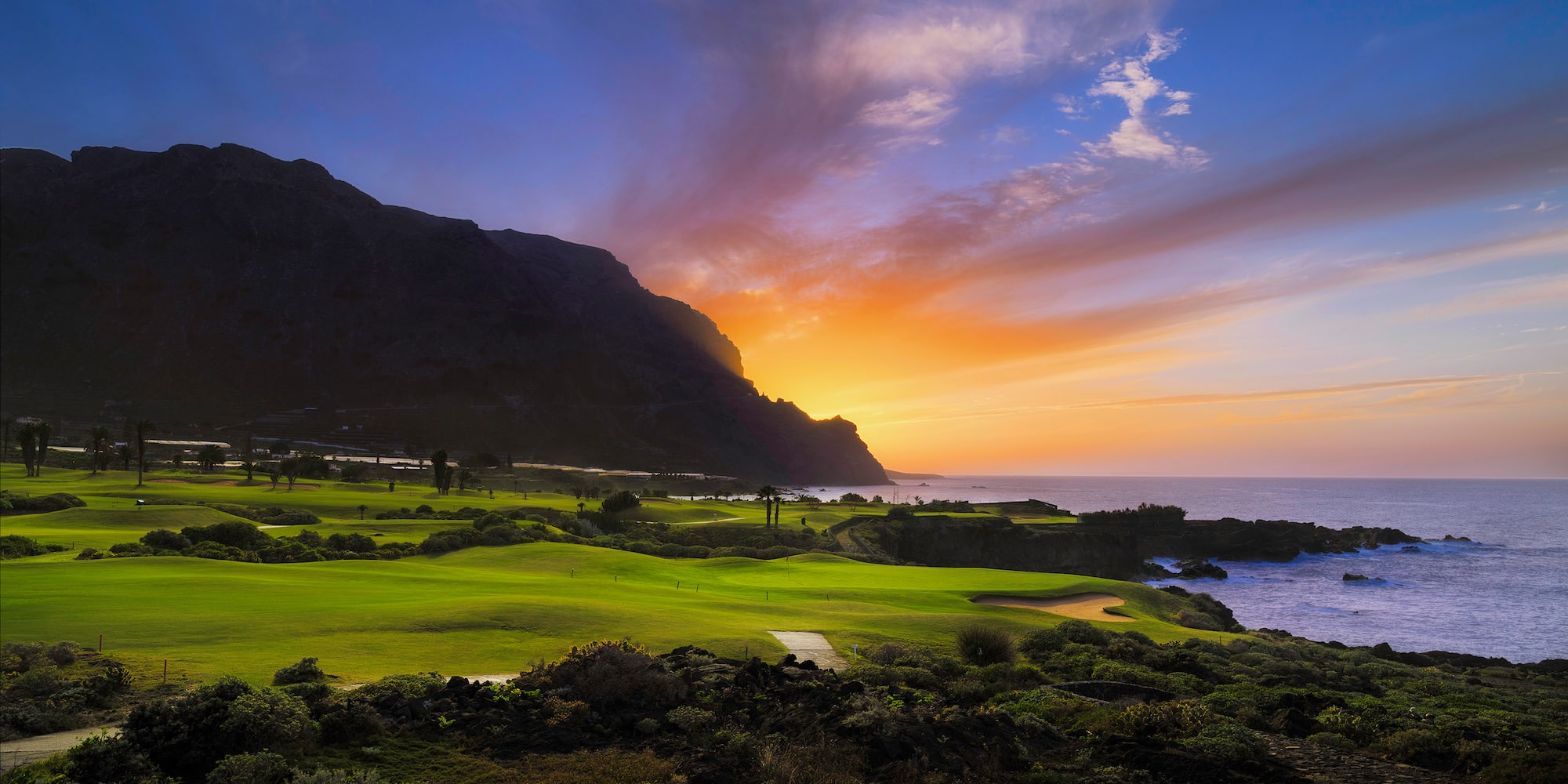 Lush green golf course by the ocean at sunset, with a mountain backdrop.