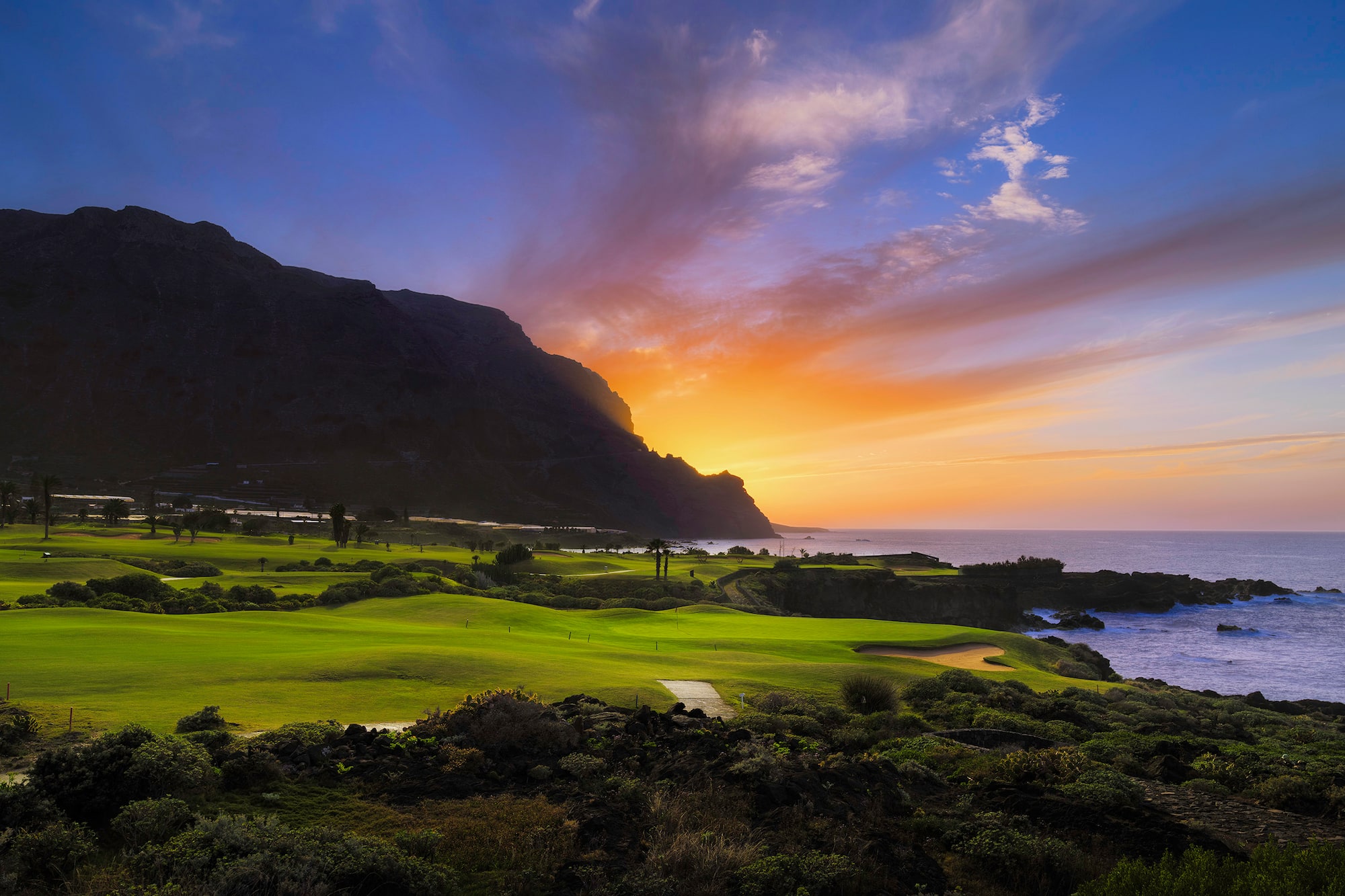 Lush green golf course by the ocean at sunset, with a mountain backdrop.