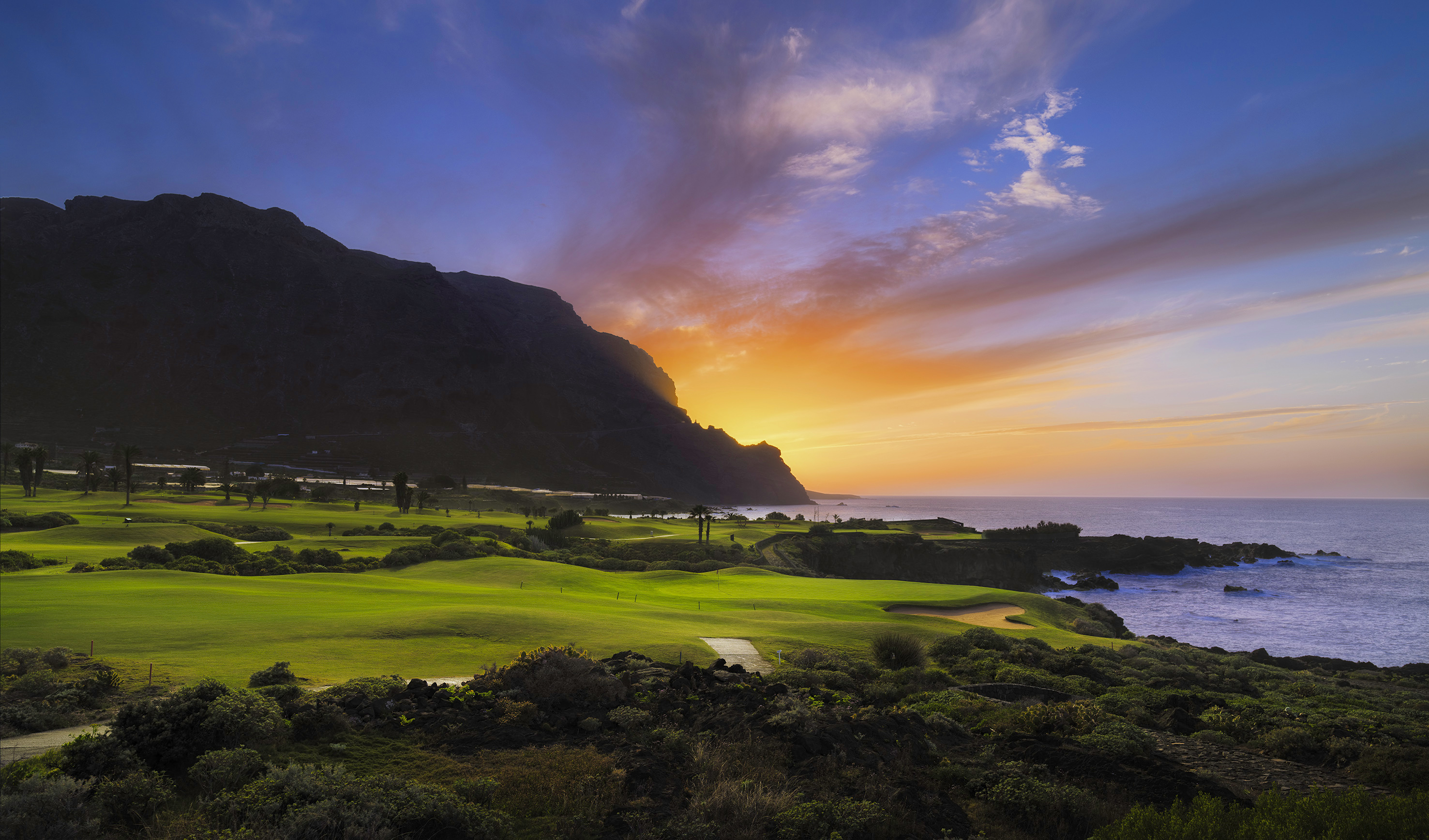 Lush green golf course by the ocean at sunset, with a mountain backdrop.