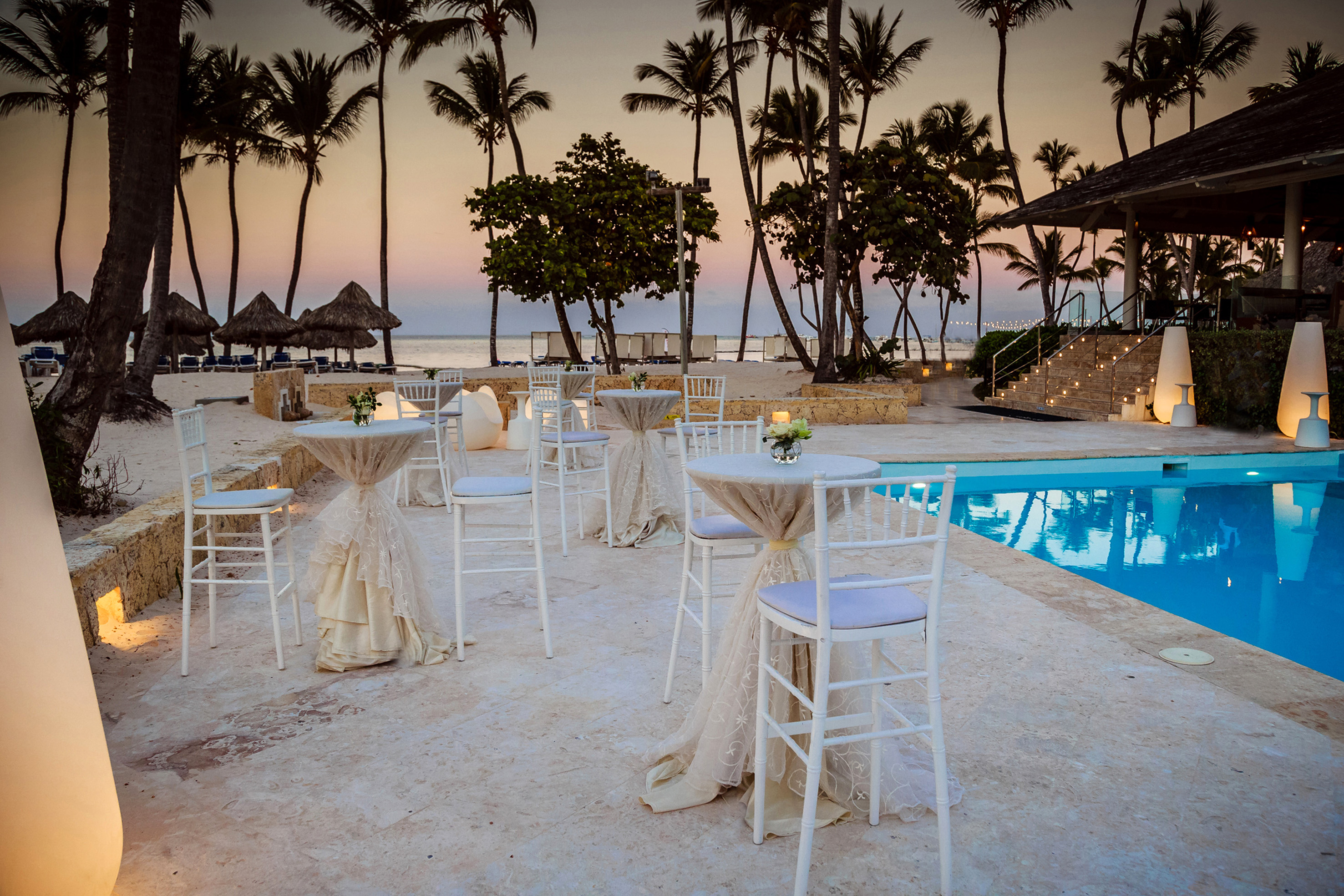 Tropical resort poolside event setup with white tables, palm trees, and sunset.