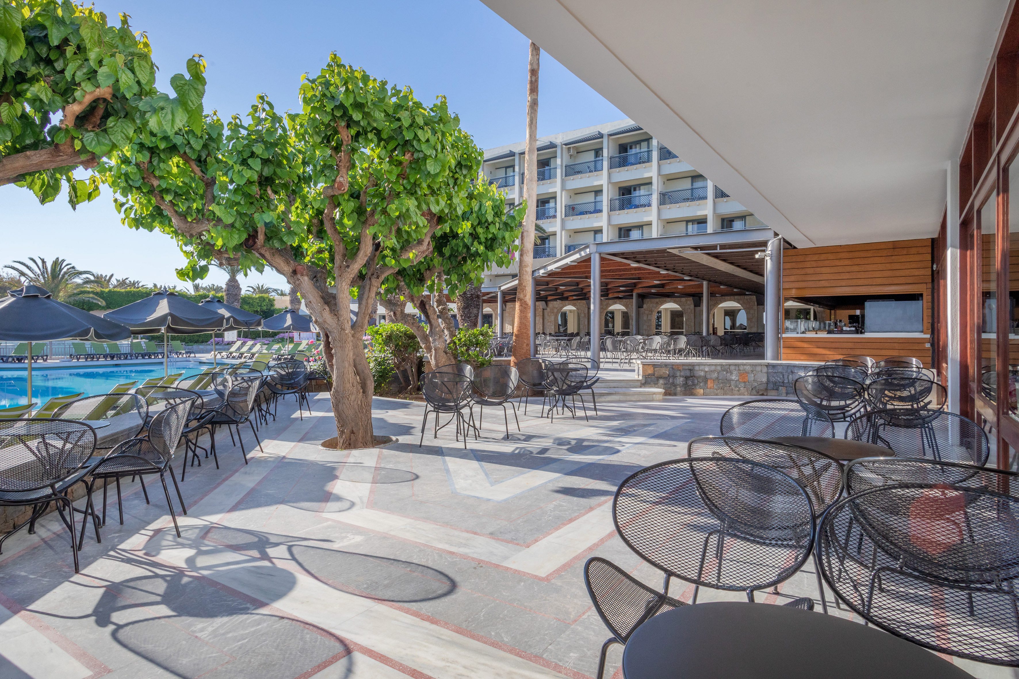 Sunny hotel terrace with cafe seating, swimming pool, green trees, and hotel building.