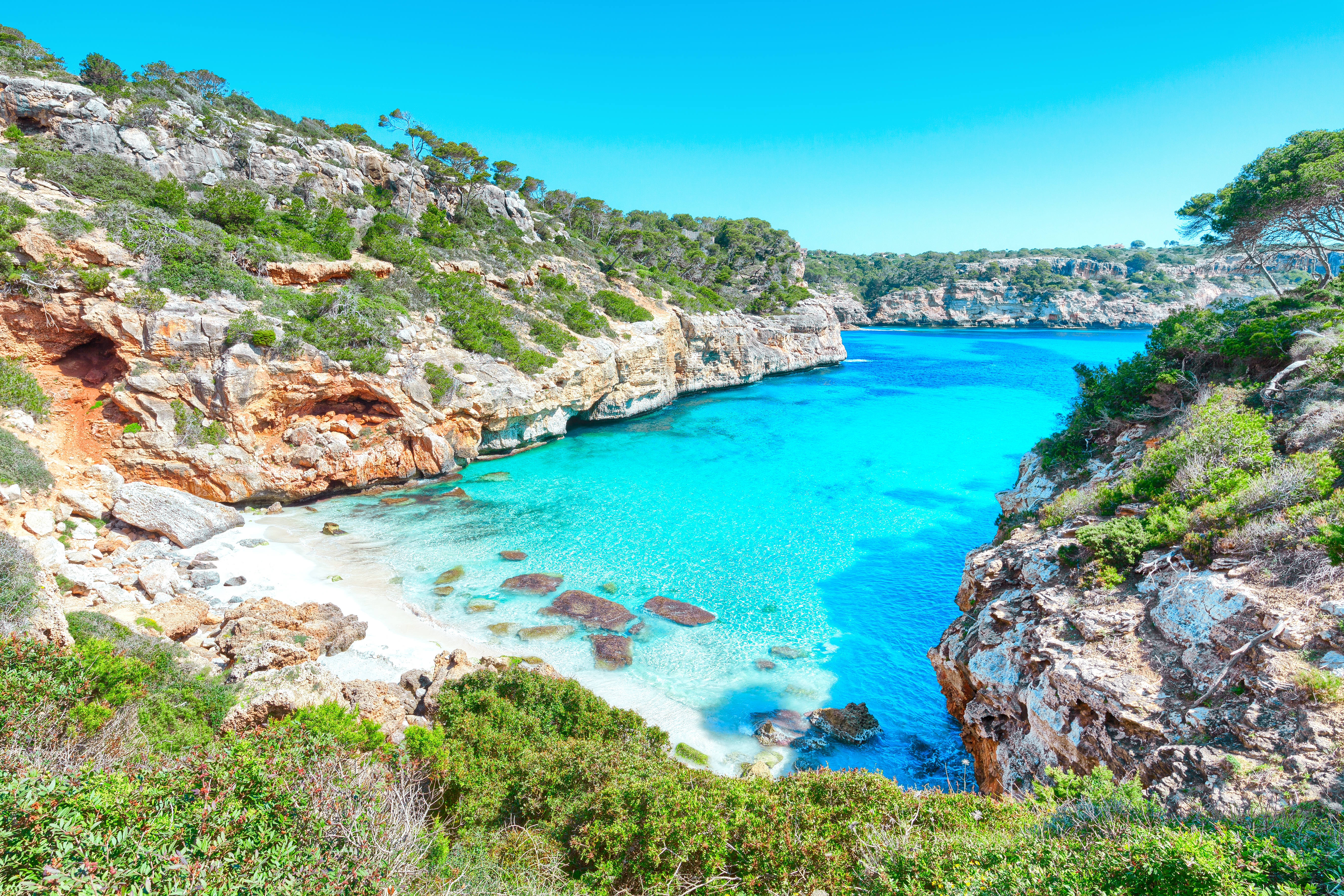 Mallorca cove: turquoise water, white sand beach, lush green cliffs.