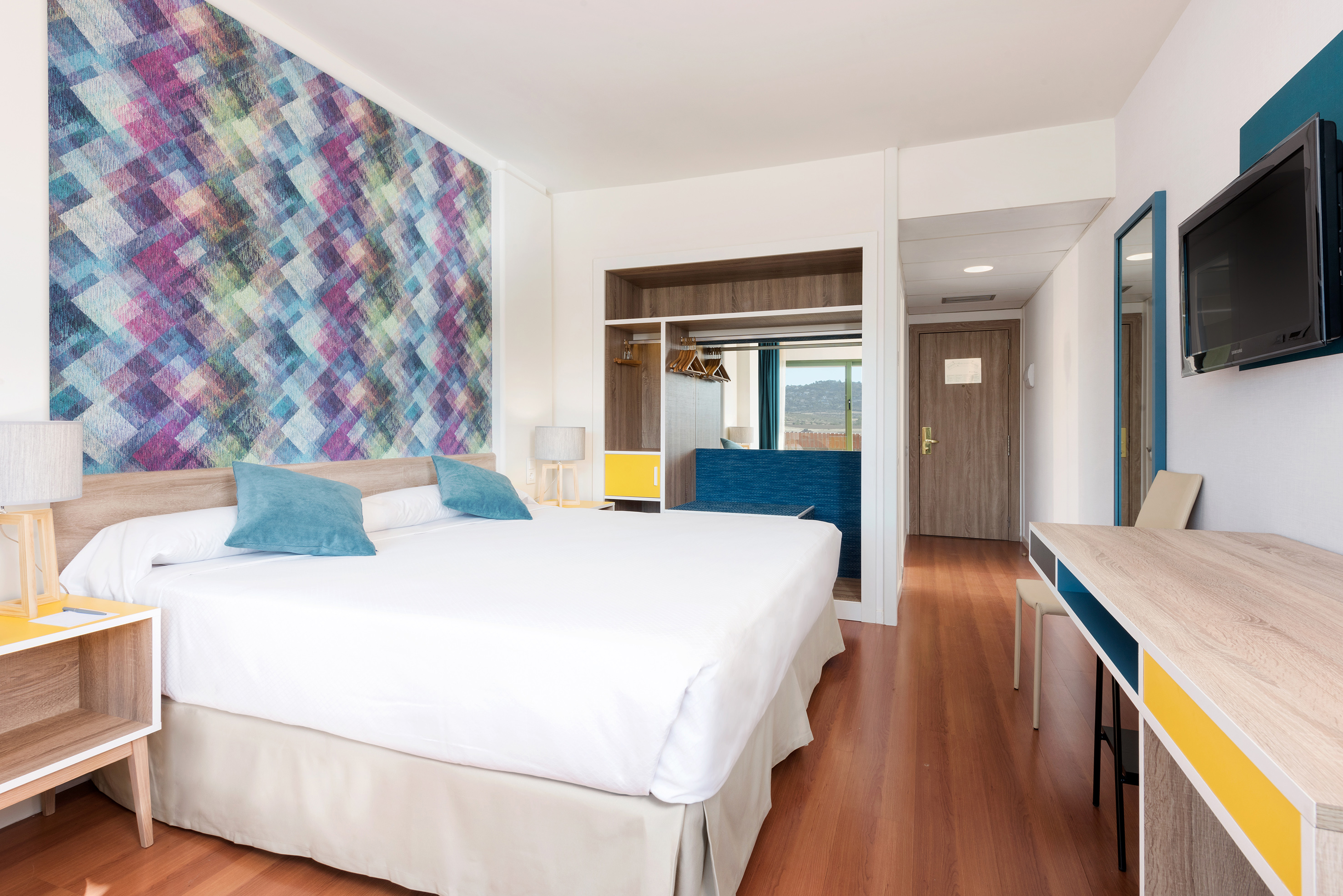Modern hotel room with large bed, vibrant geometric wall, and wooden floor.