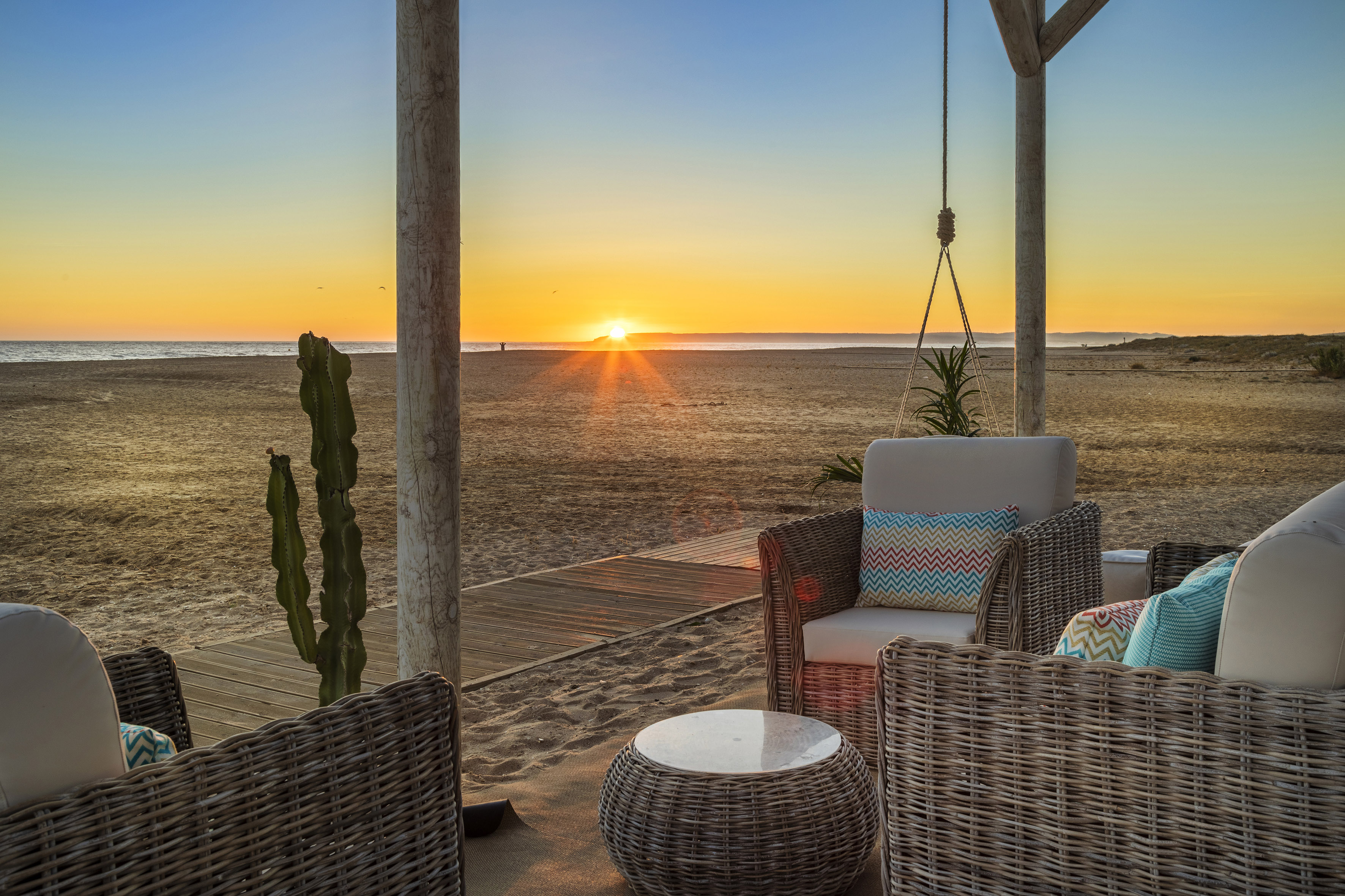 Sunset on sandy beach from a lounge patio with wicker chairs and cactus.