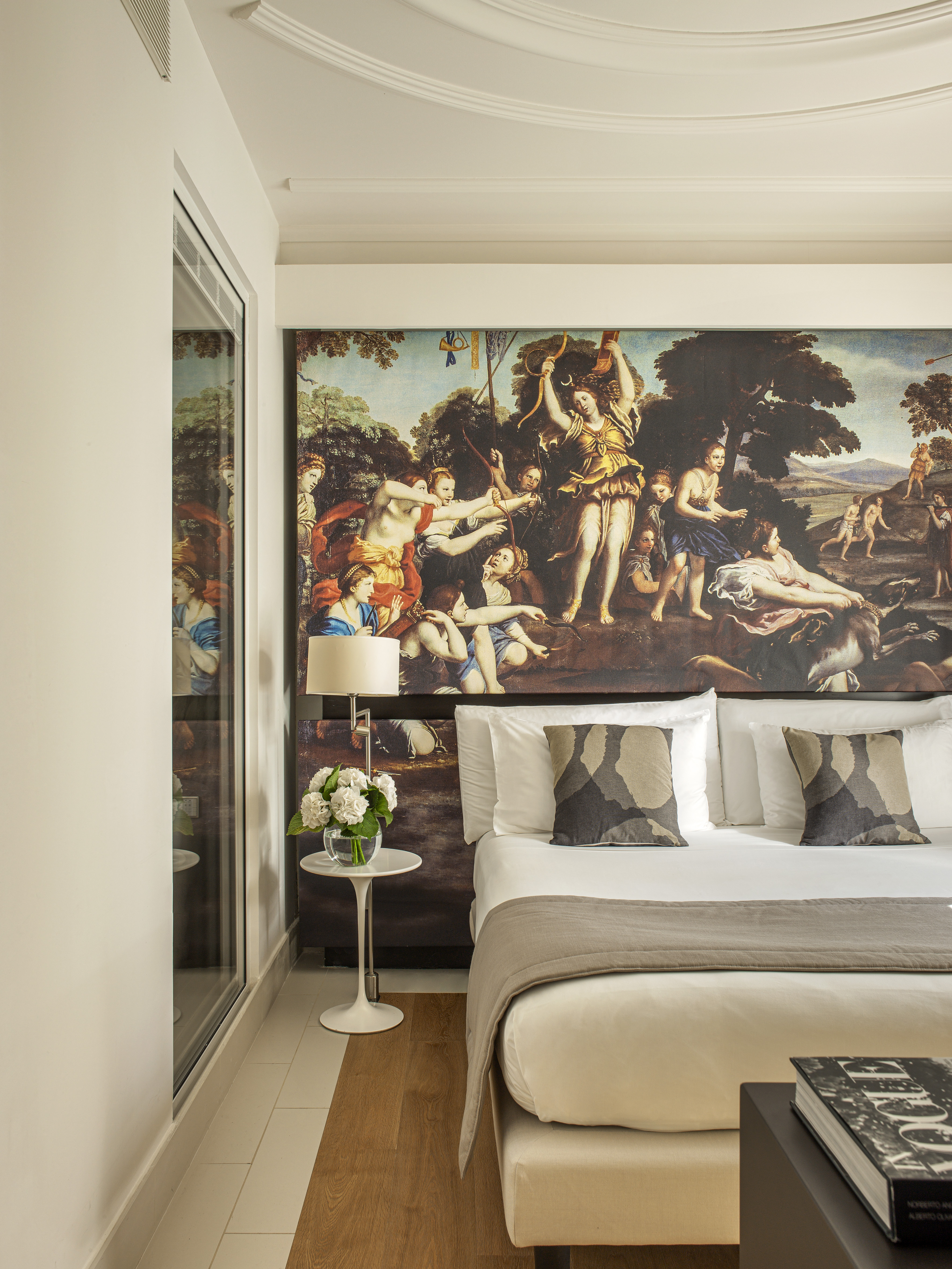 Hotel room with large classical mural above bed, crisp white bedding, and modern side table.