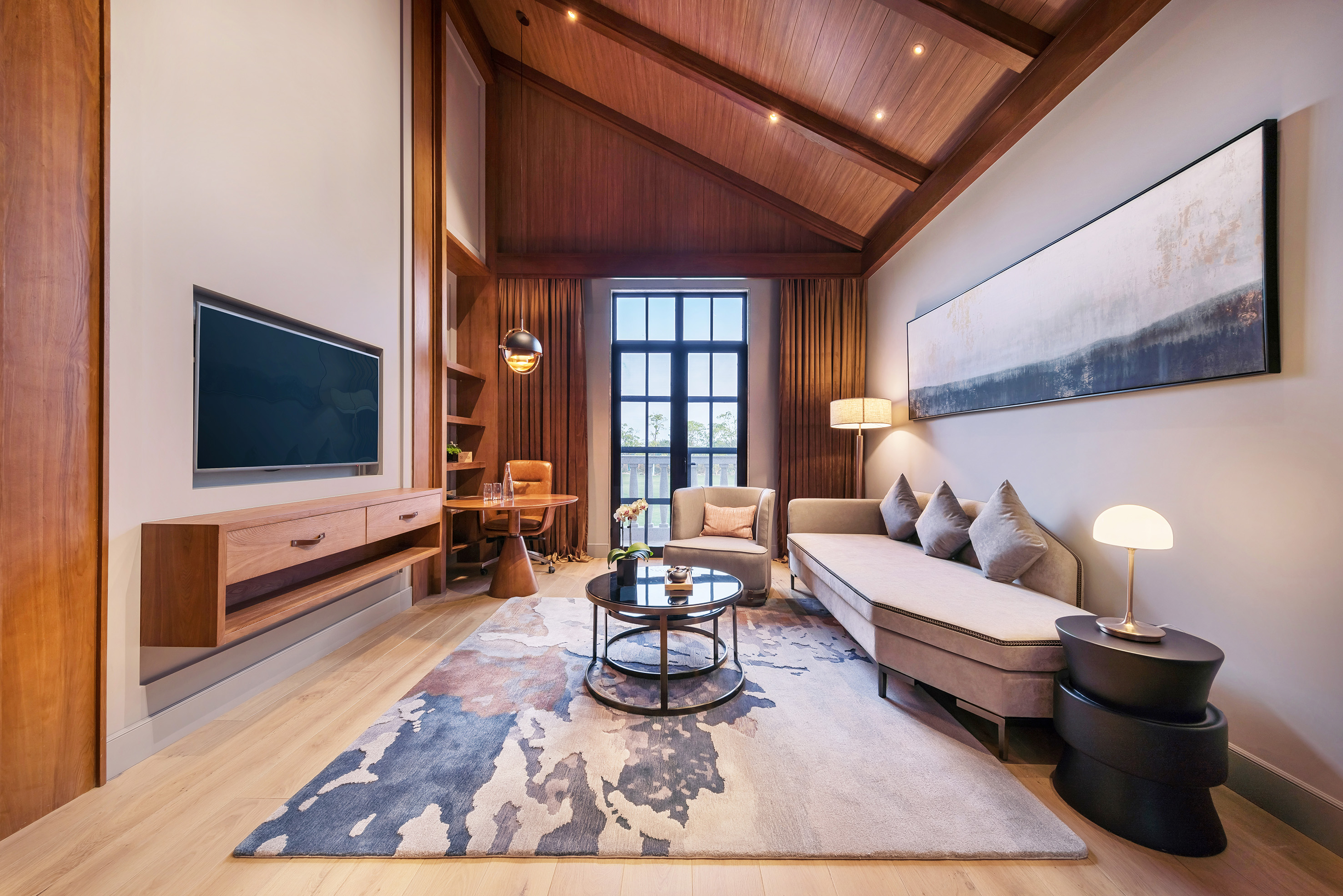 Modern hotel suite with vaulted wood ceiling, sofa, TV, desk, and abstract rug.