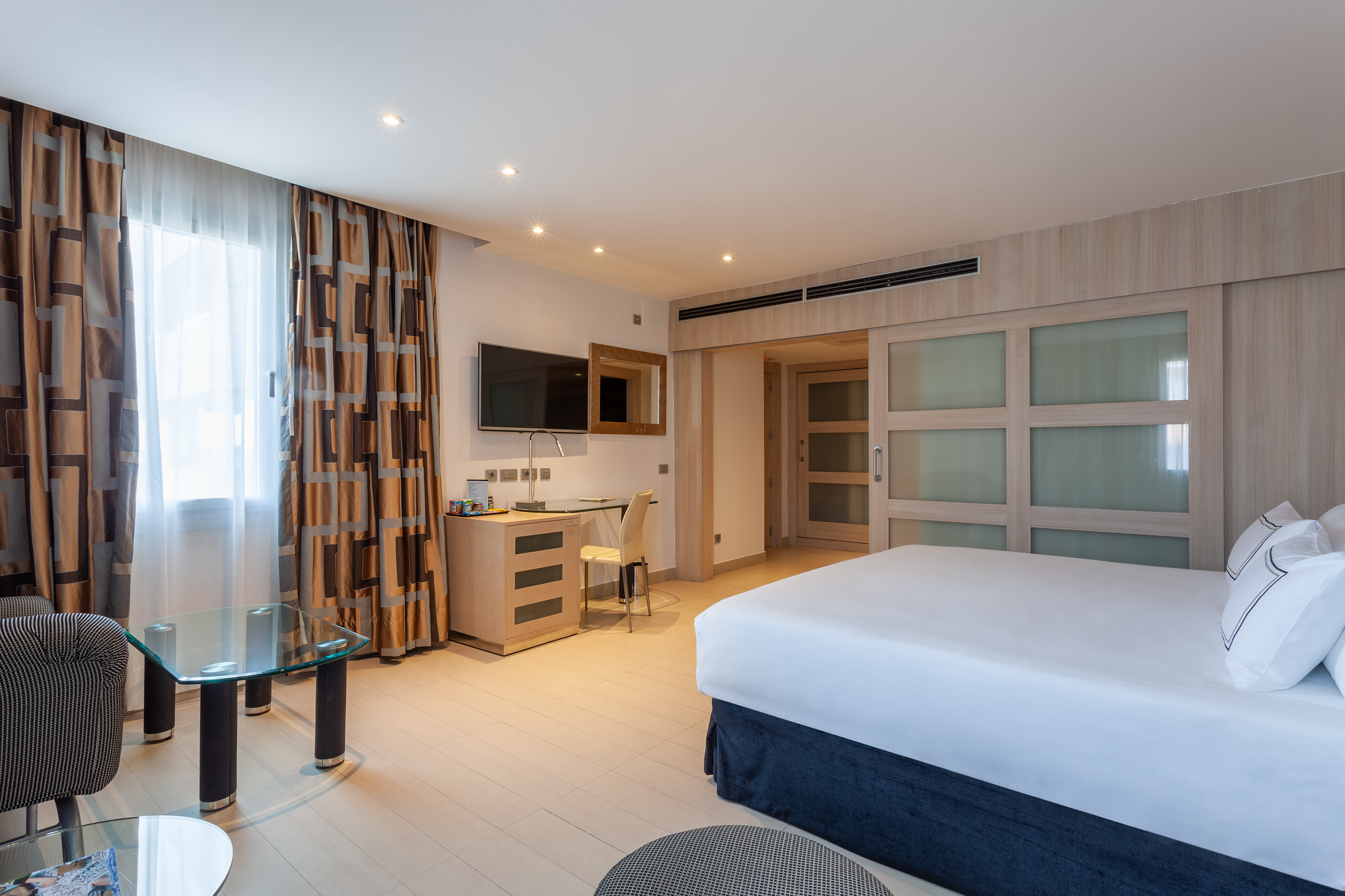 Modern hotel room with bed, desk, TV, window, and sliding wooden closet.