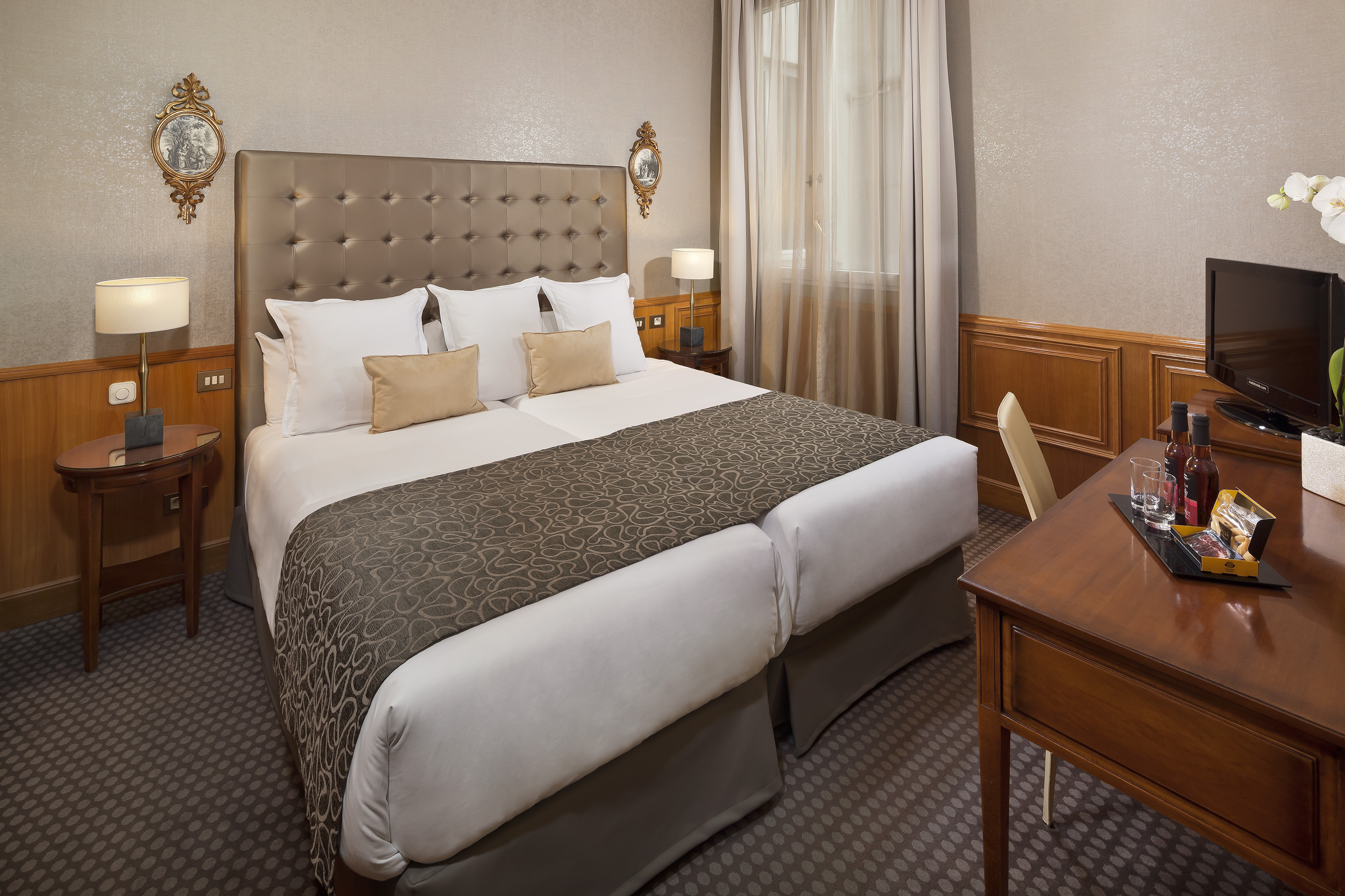 Elegant hotel room with twin beds, tufted headboard, and wooden desk with minibar.