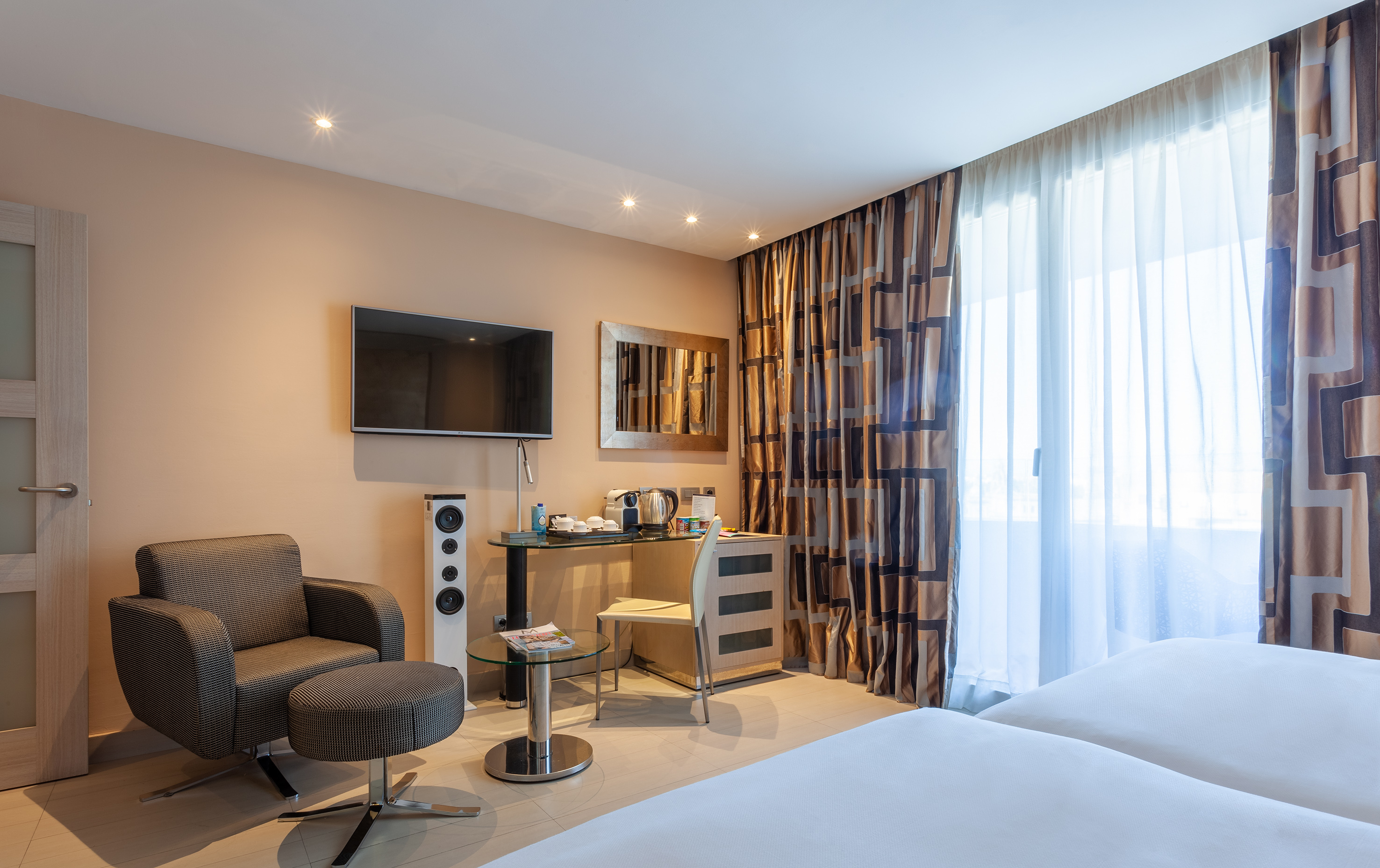 Modern hotel room with armchair, wall-mounted TV, desk, and large window with curtains.