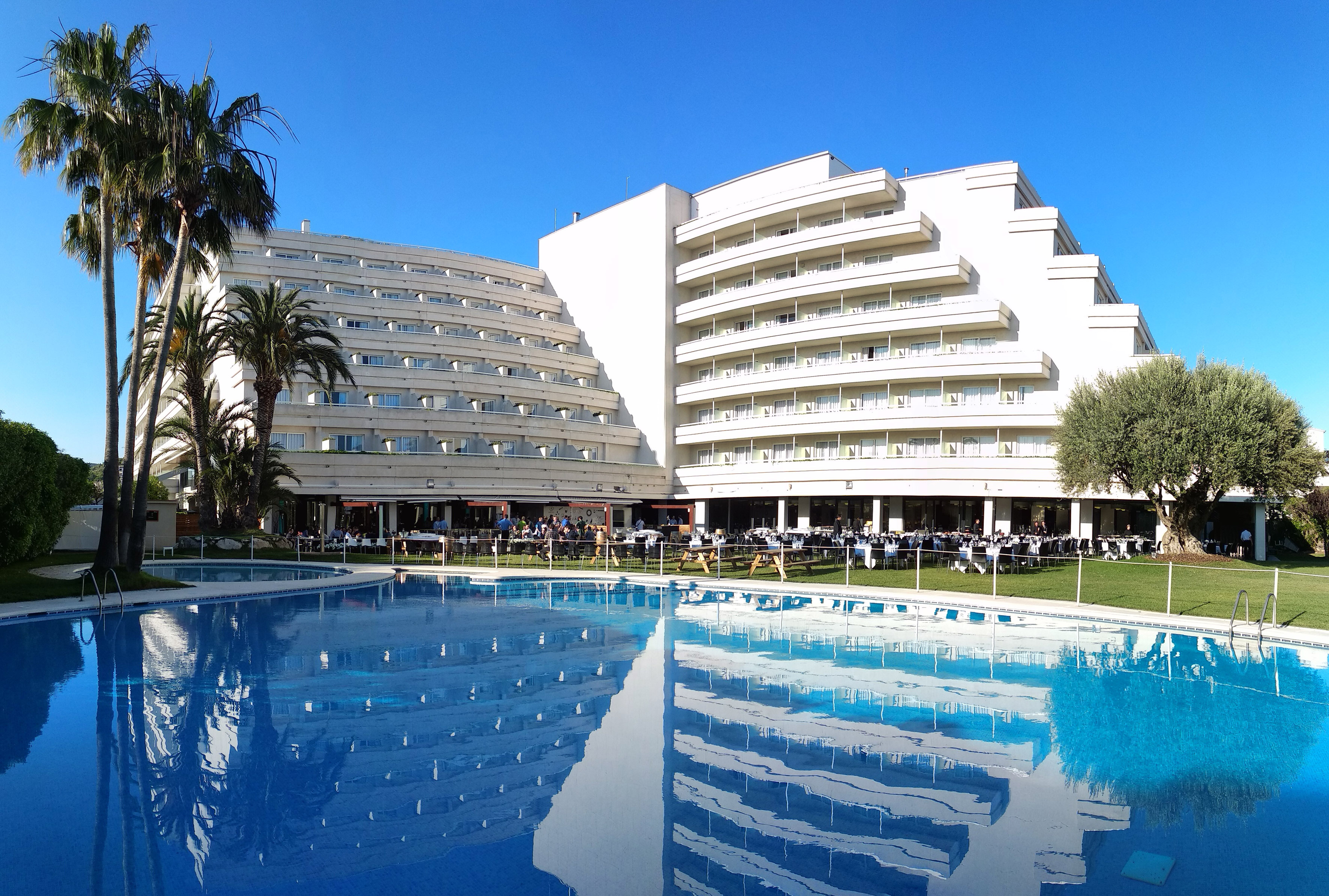 White hotel with large swimming pool, palm trees, and outdoor dining area.