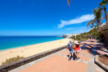 a couple of people walking on a sidewalk with a beach and blue water