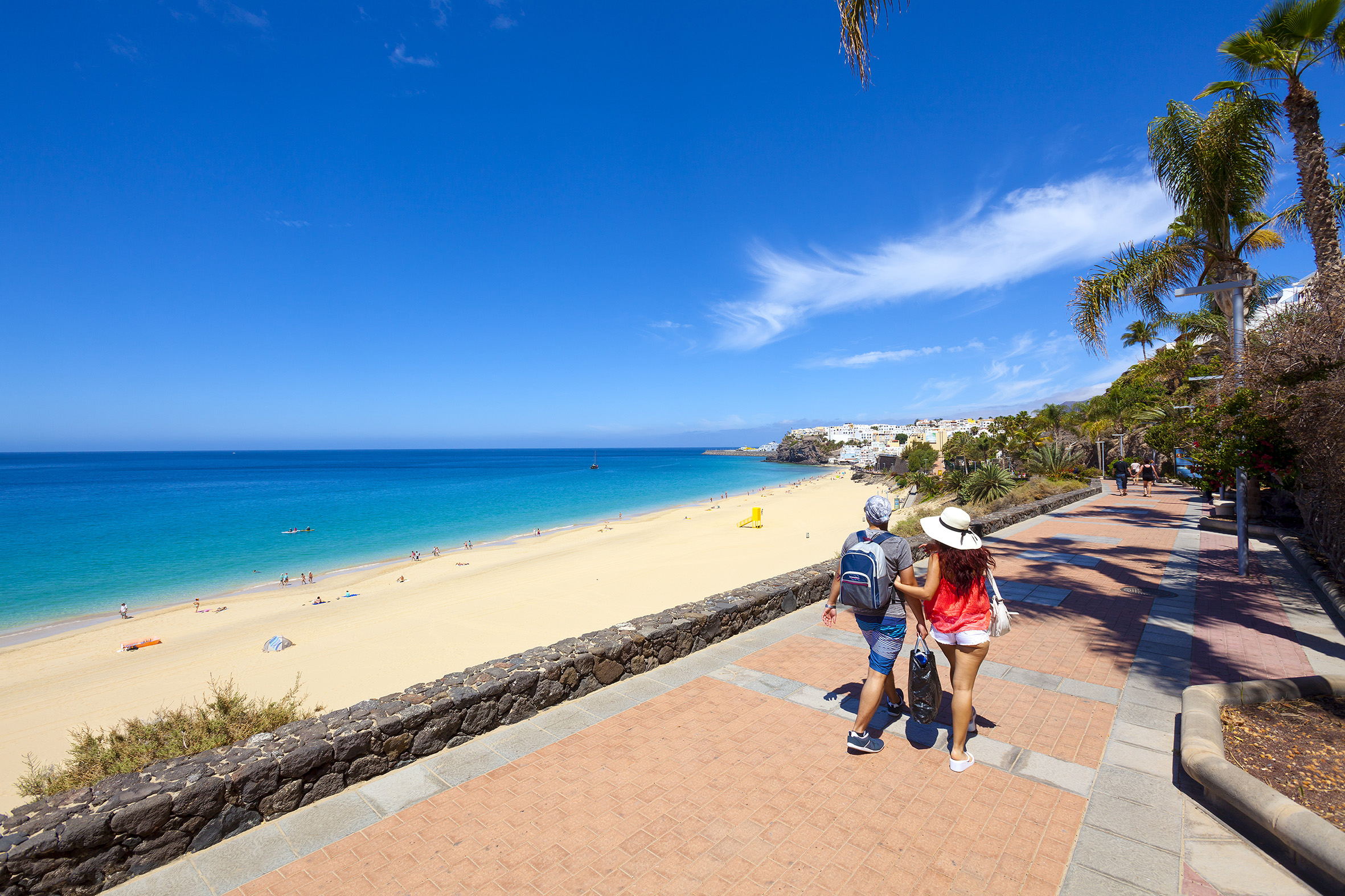 a couple of people walking on a sidewalk with a beach and blue water