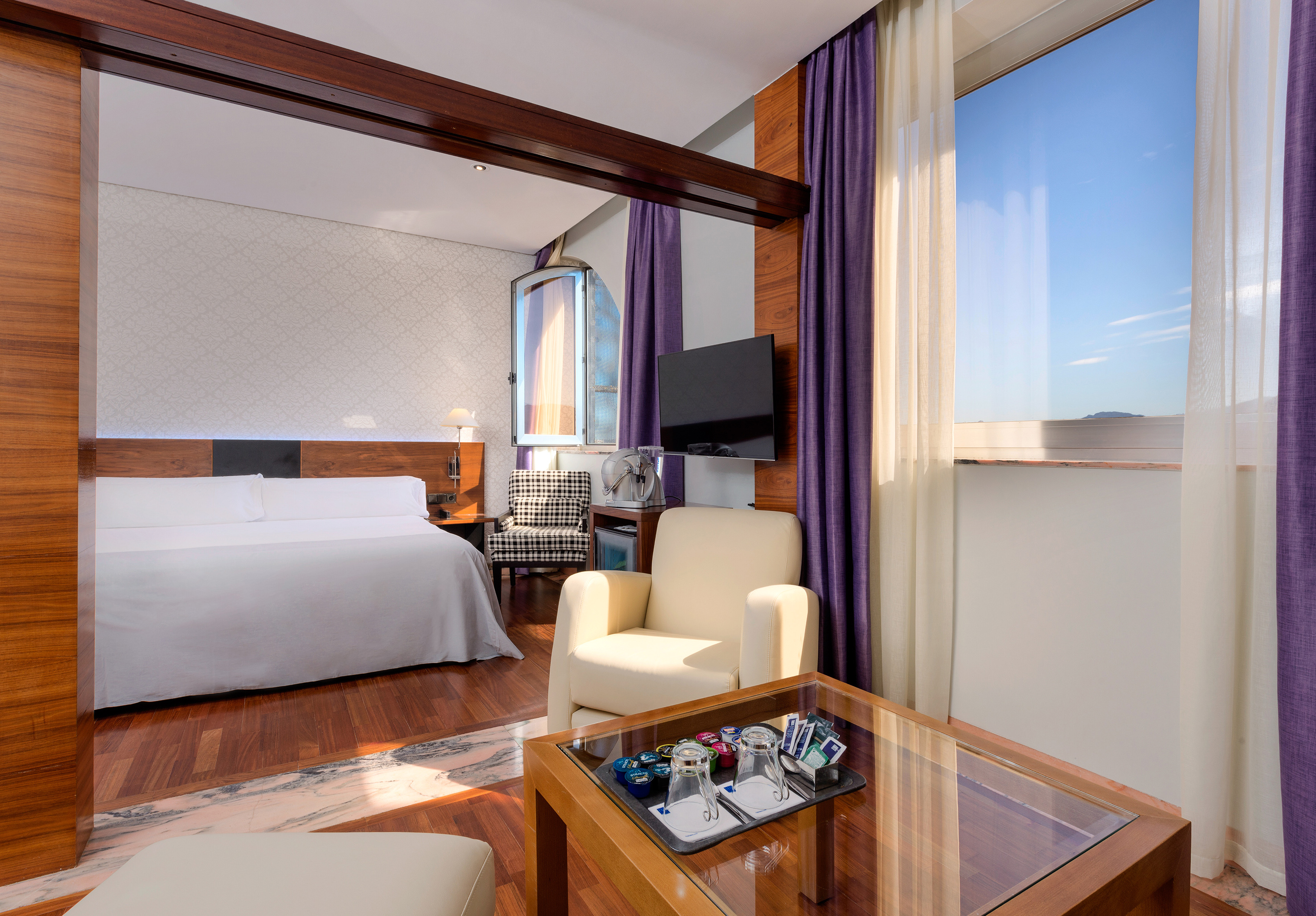 Luxury hotel suite with king bed, wooden floors, purple curtains, and large window.