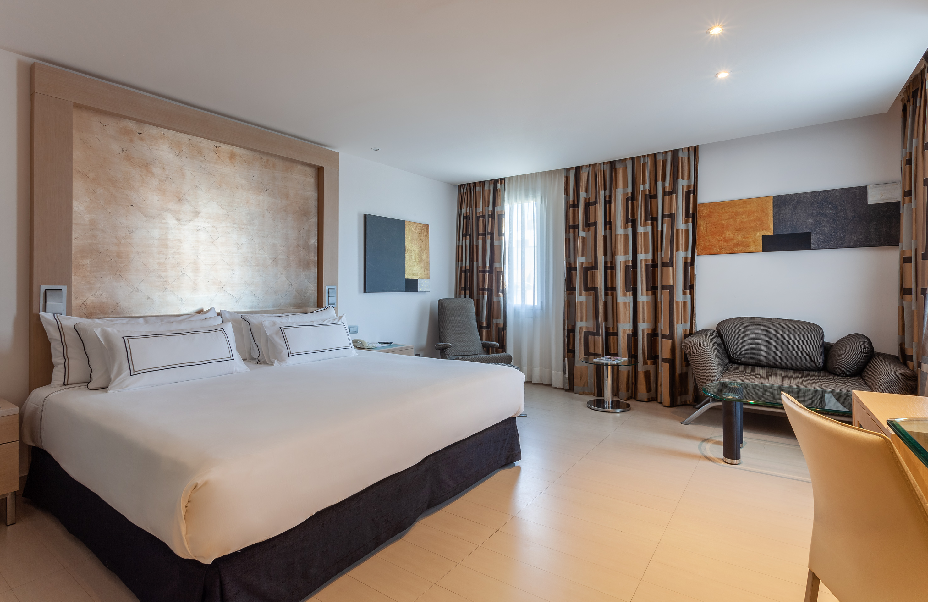 Modern hotel bedroom with king bed, sofa, lounge area, and stylish decor.