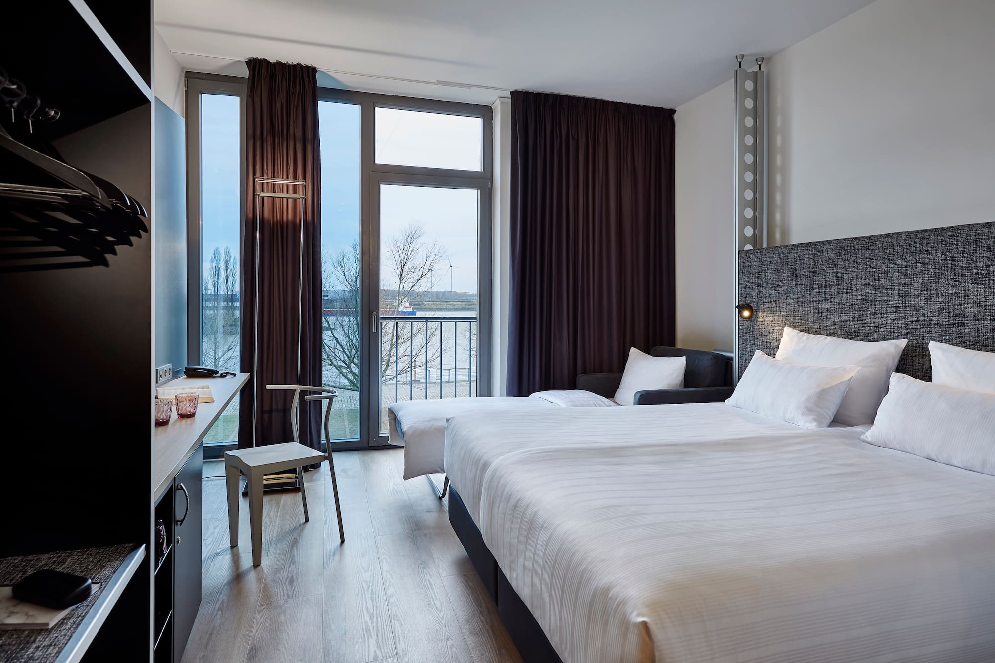 Modern hotel room with twin beds, desk, waterfront view, and dark curtains.