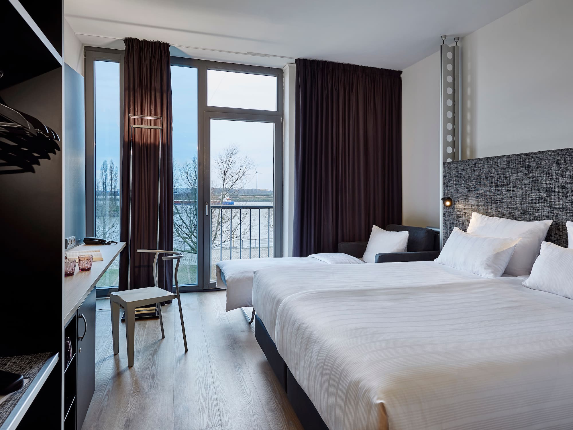 Modern hotel room with twin beds, desk, waterfront view, and dark curtains.