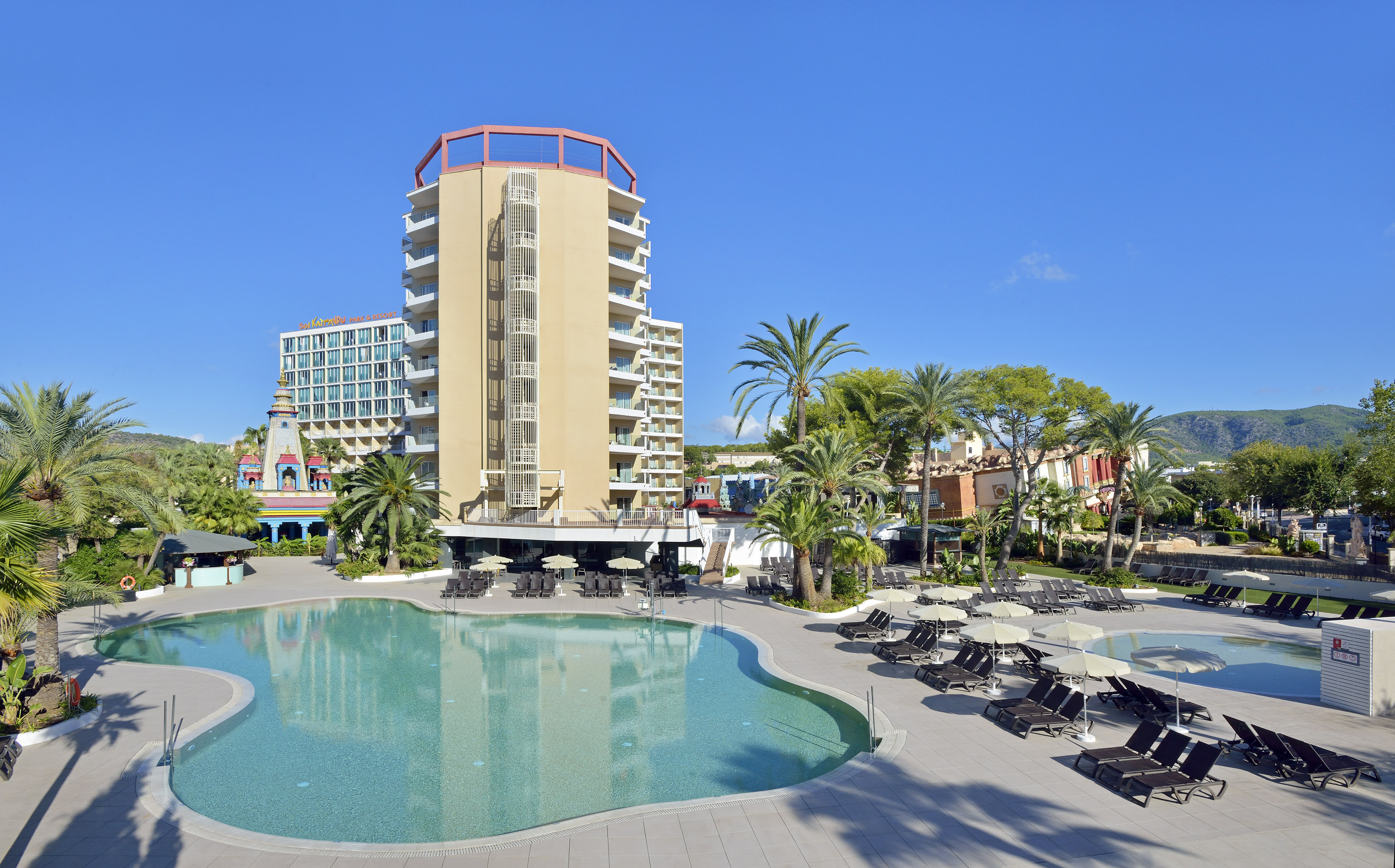 Sunny resort hotel with large outdoor pool, palm trees, and sun loungers.