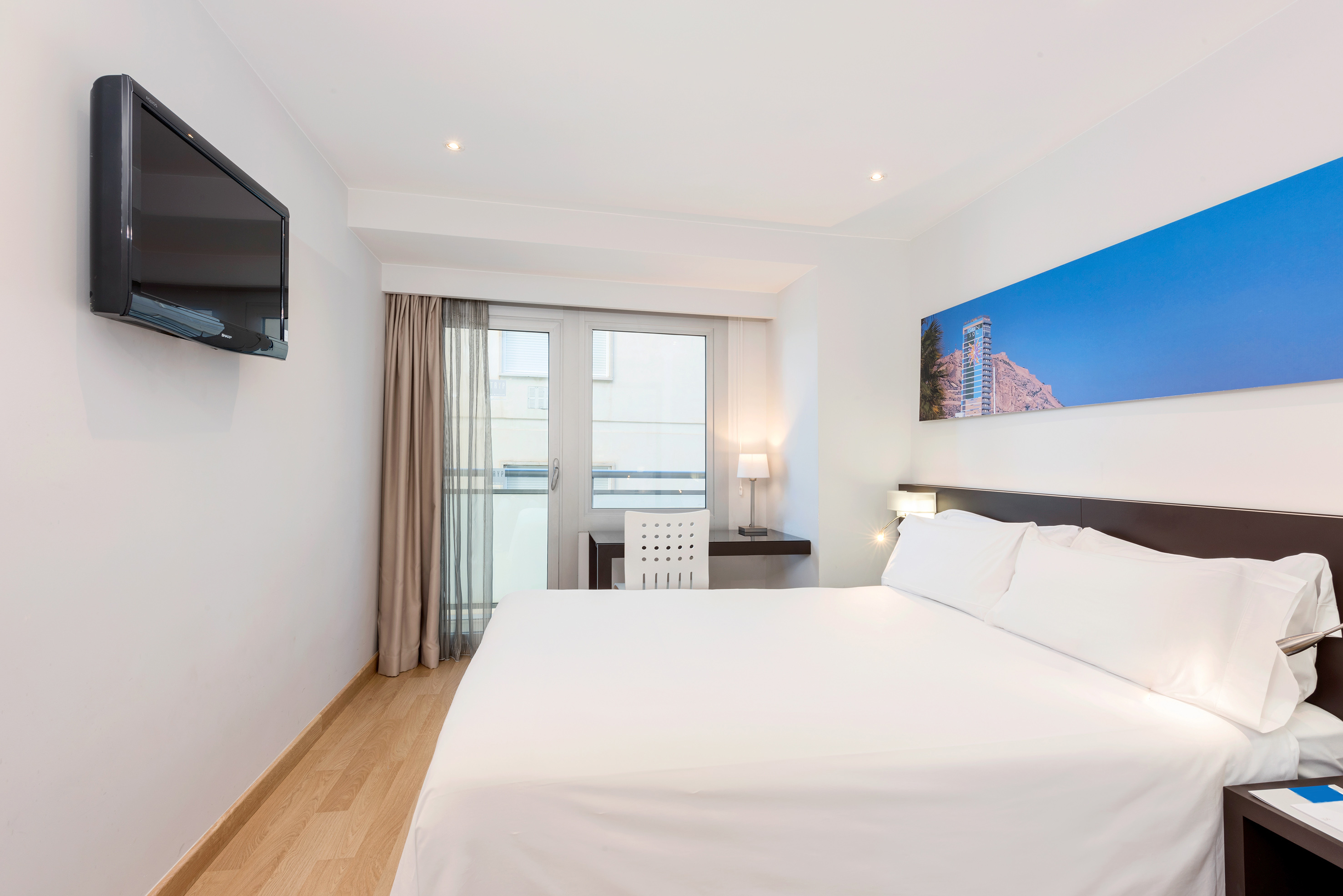 Modern hotel room with white bed, flat-screen TV, desk, and balcony door.