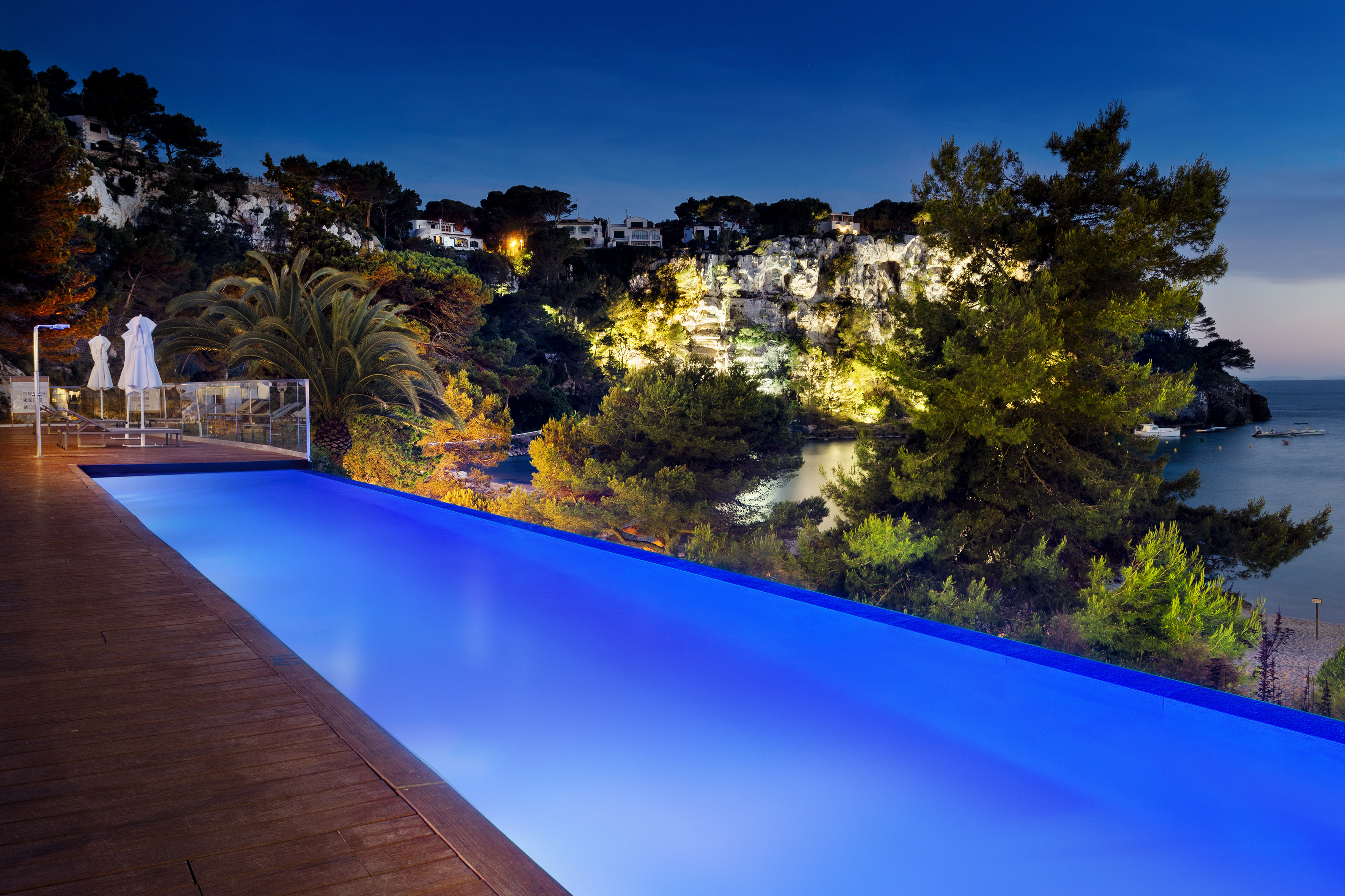 Luxury hotel infinity pool at night overlooking illuminated cliffs and sea.